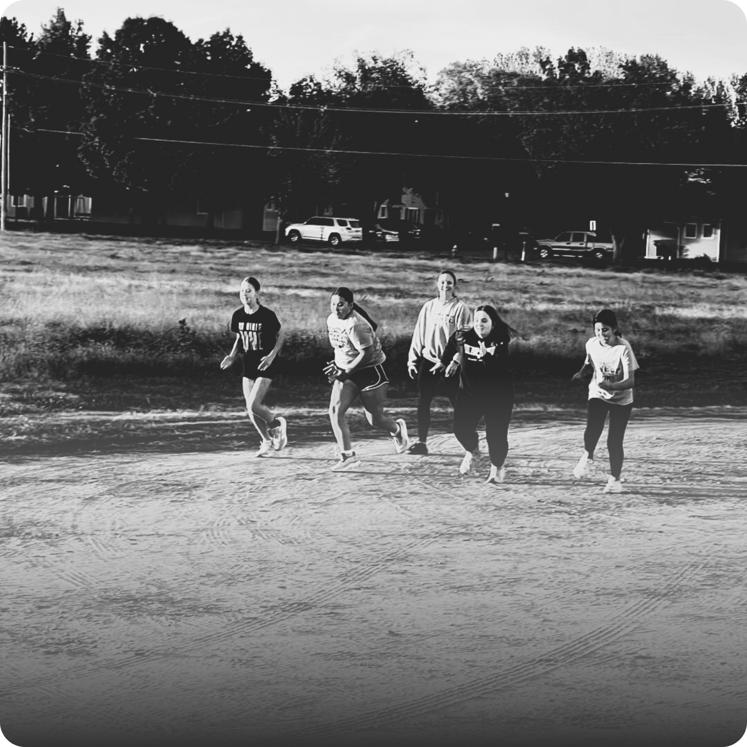 A group of five children running or jogging outdoors on a dirt surface, with a grassy area and parked cars in the background, in black and white.
