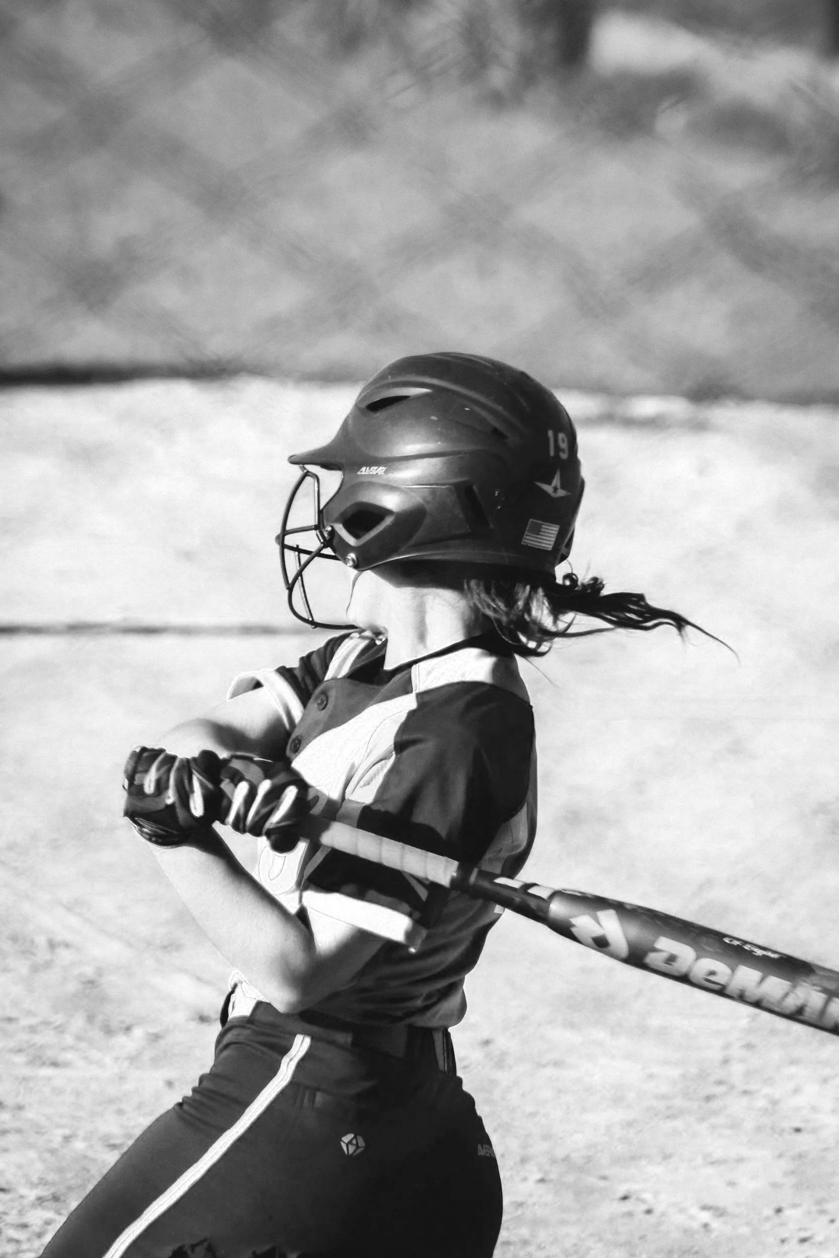 A female baseball player in a helmet, jersey, and gloves, swinging a bat on a baseball field.