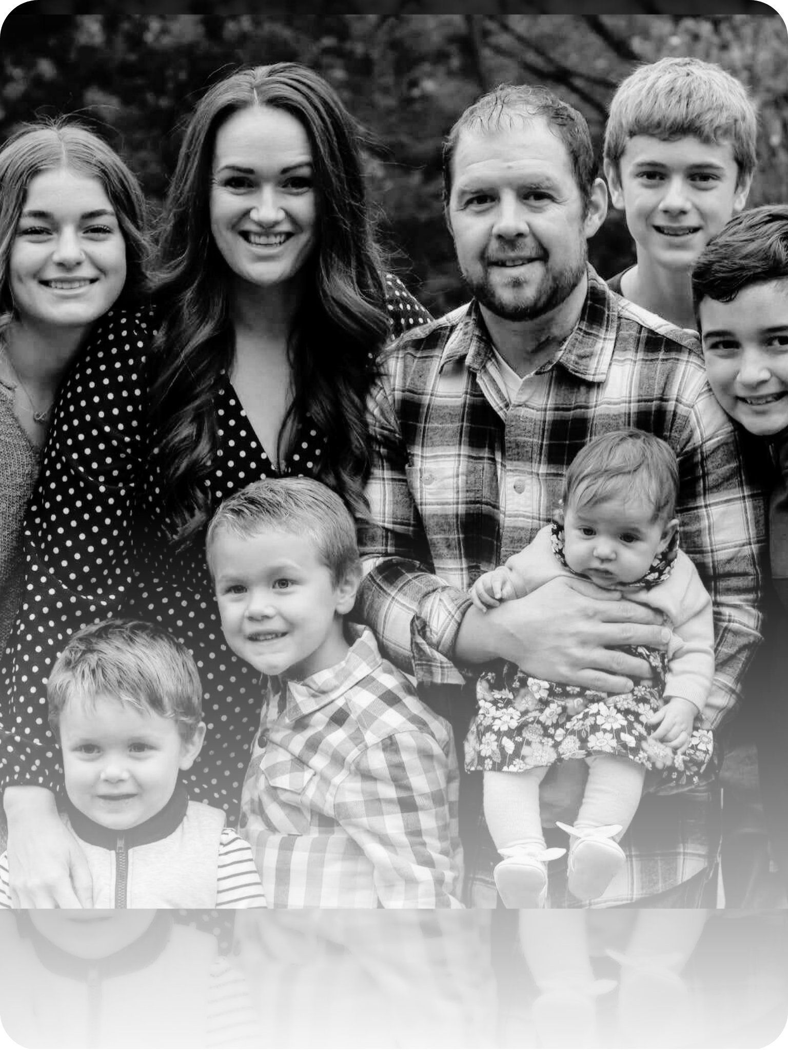 A black and white photo of a family of seven outdoors, including three adult women and men, and four children, with trees in the background.