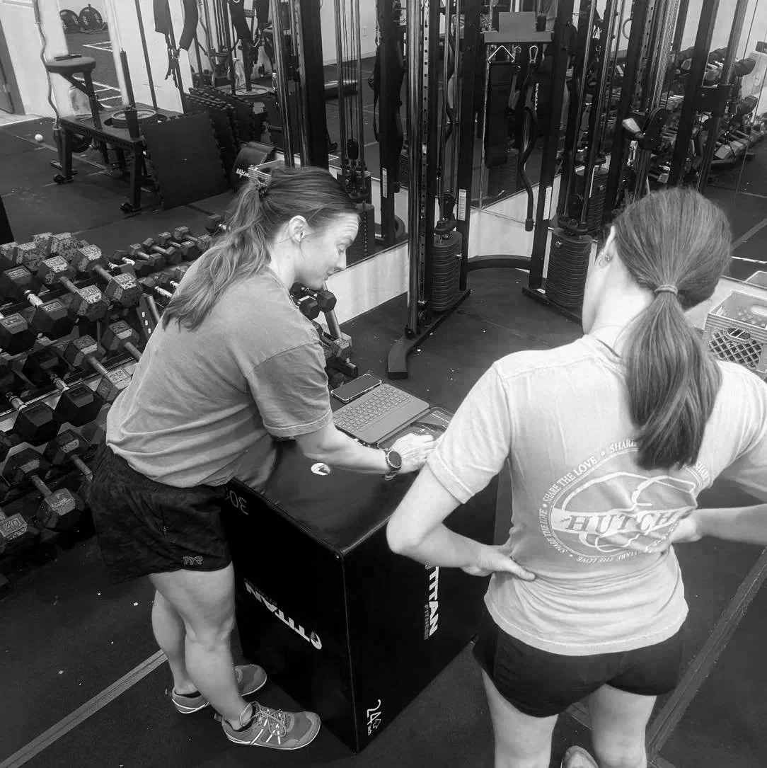 Two women in a gym looking at a tablet on a plyometric box. One woman is leaning over the box, and the other is standing with her hands on her hips. There are dumbbells and gym equipment in the background.