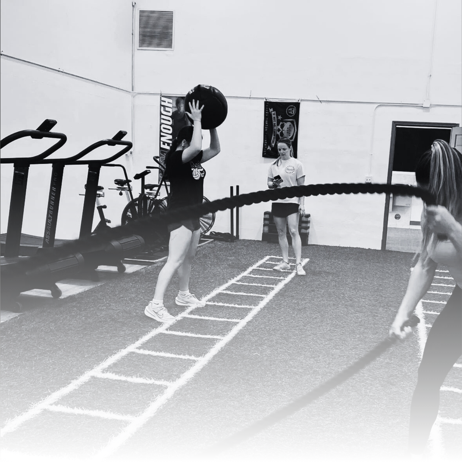 Women working out with medicine balls and ropes in a gym, with fitness equipment and motivational banners visible.