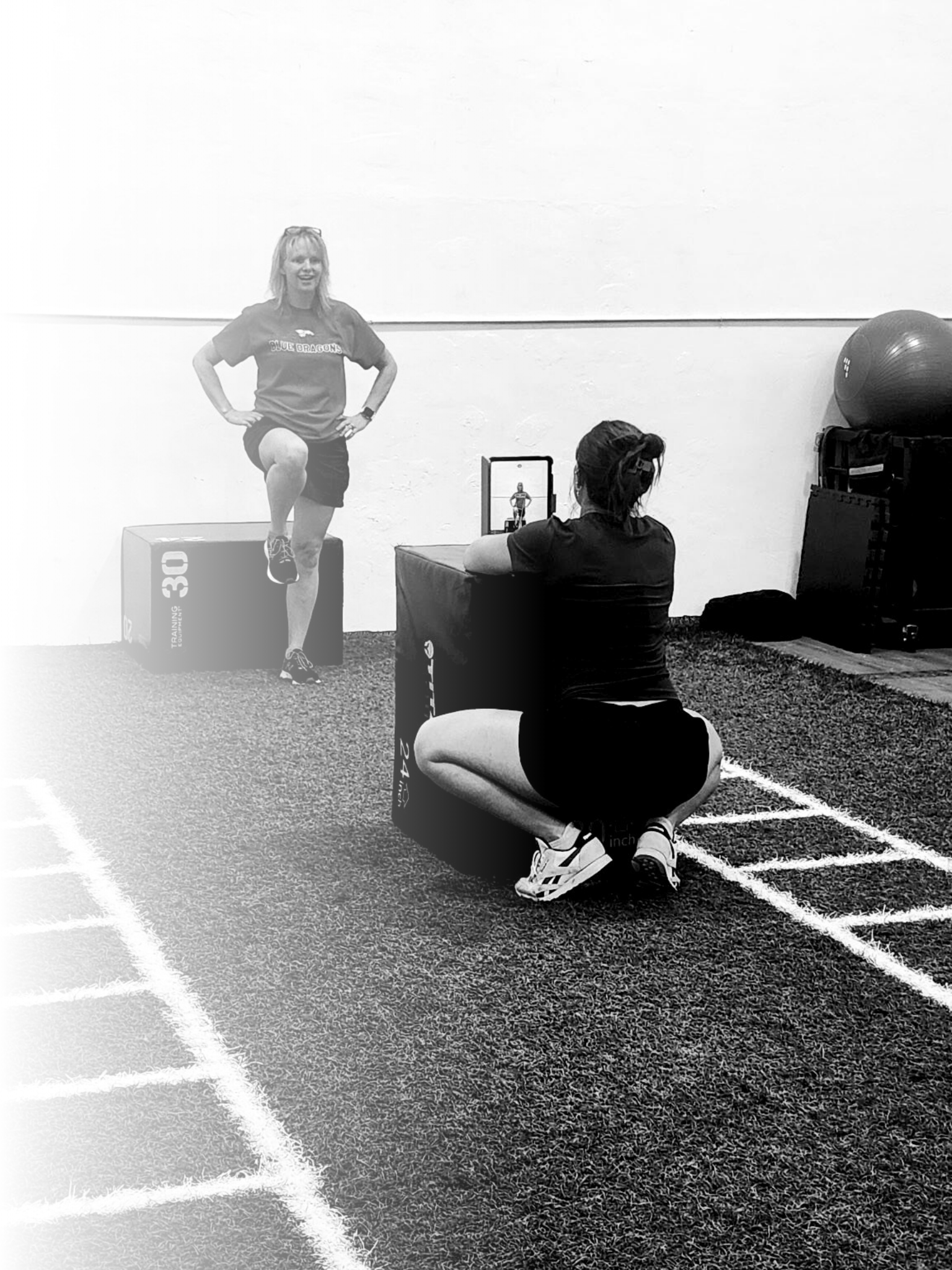 A woman exercises in a gym, lifting her knee onto a plyometric box while another woman kneels on the ground taking a photo or video with her phone, with fitness equipment visible in the background.
