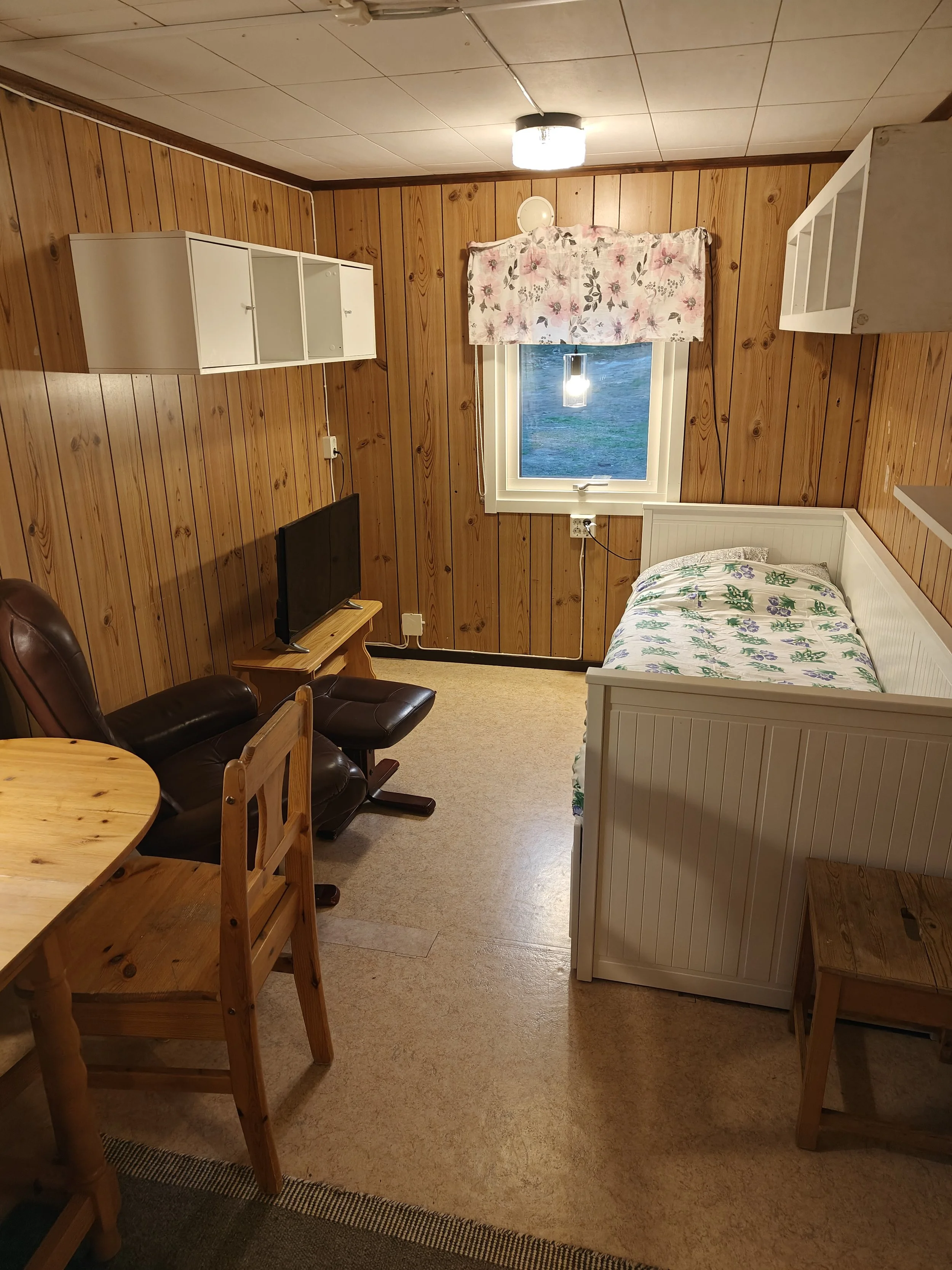 A cozy room with wooden panel walls and a single bed with floral bedding, a small window with floral curtains, a TV on a small wooden stand, a leather armchair with an ottoman, and a small wooden table.