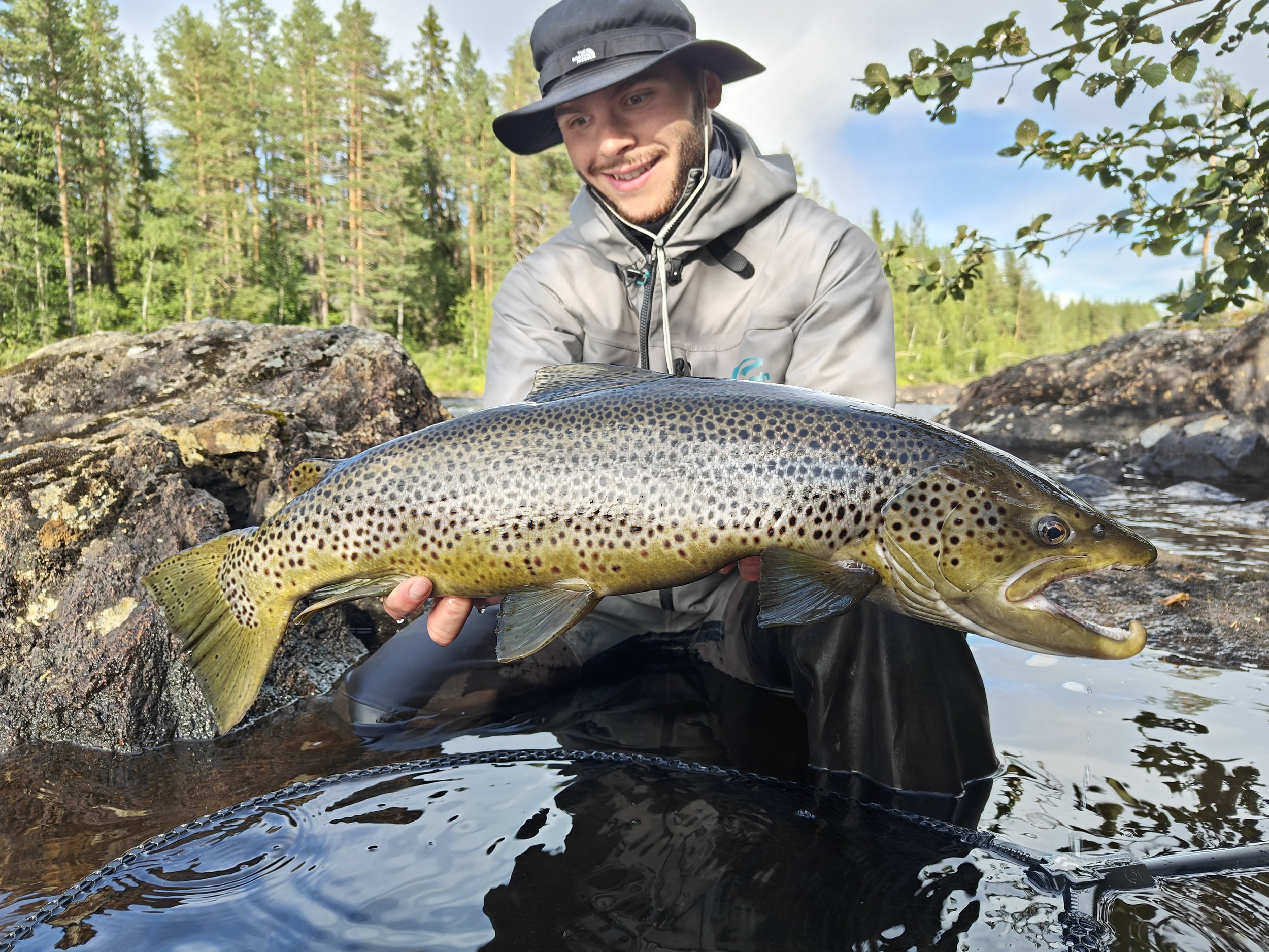 A man in outdoor gear holding a large sea trout in a river with rocks and trees in the background.