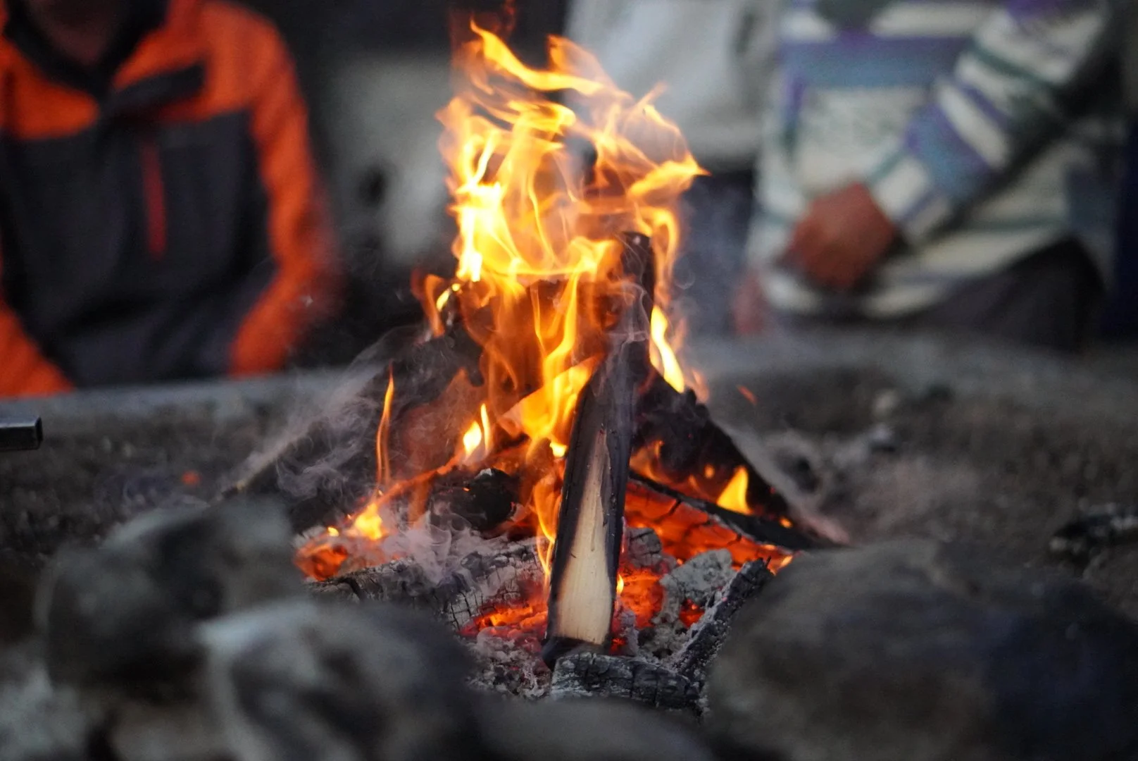Close-up of a campfire with bright orange flames burning wood and glowing embers on the ground, with blurred people in outerwear visible in the background.