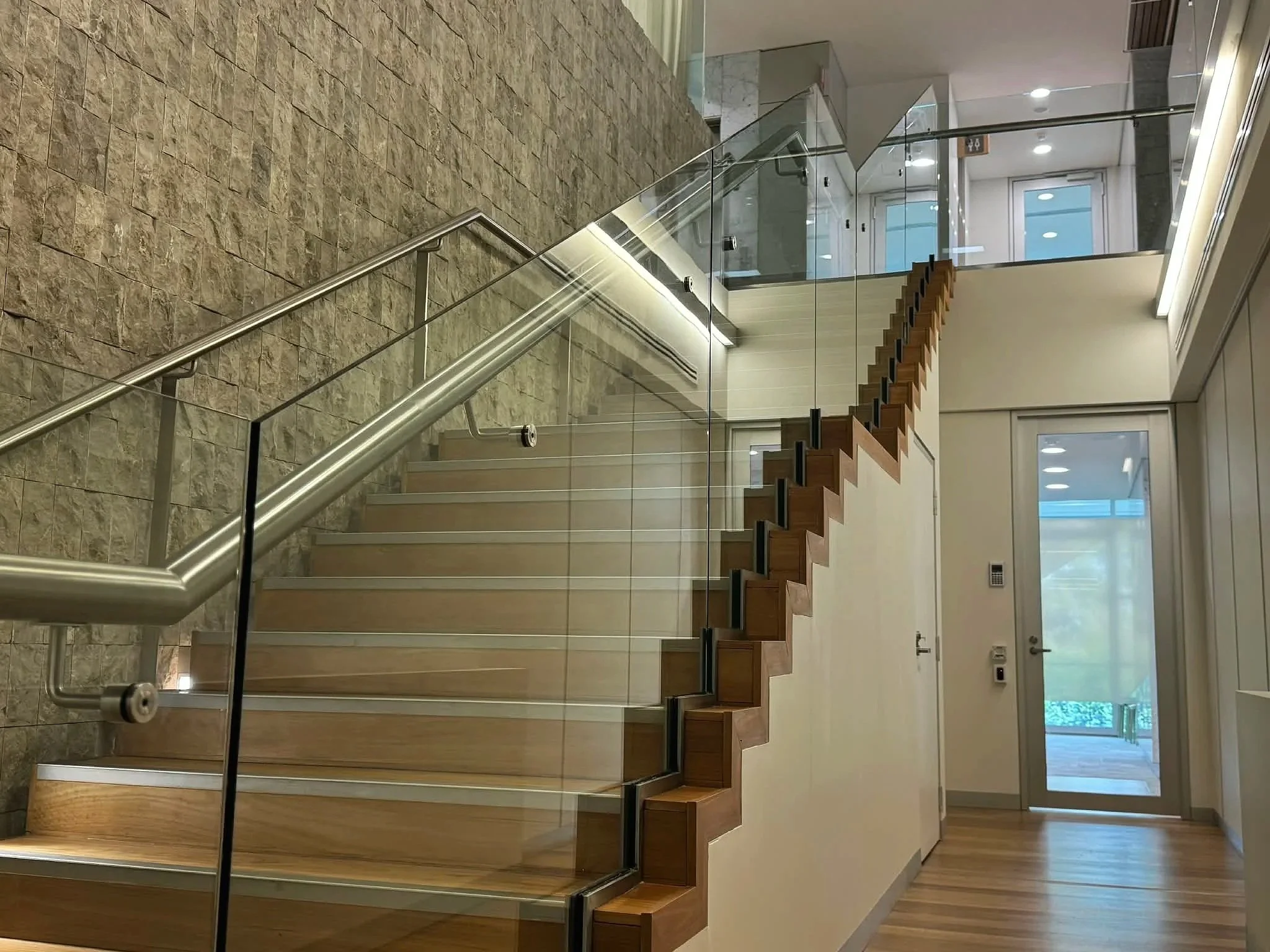 Interior of a modern building with a wooden staircase, glass railing, stone wall, and glass door leading outside.