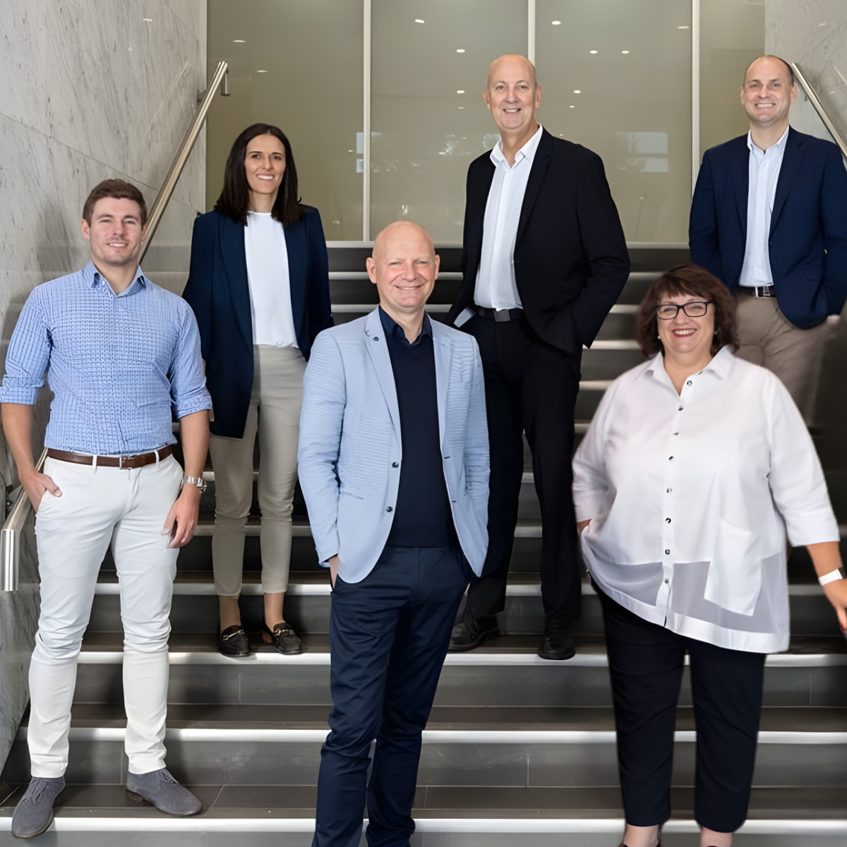 Group of seven professionally dressed people standing on stairs inside a modern office building, smiling at the camera.