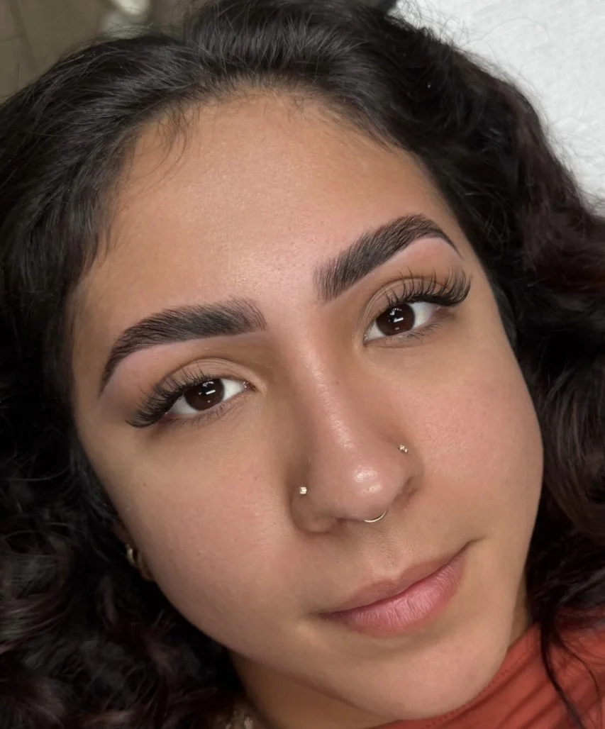 Close-up of a woman with dark curly hair, groomed eyebrows, long eyelashes, and a nose piercing with small rings.