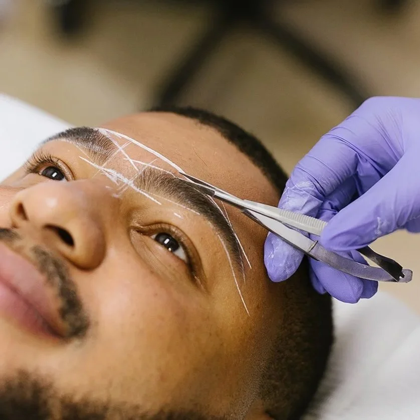 Person lying down and receiving an eyebrow tattooing procedure, with a person wearing purple gloves using surgical tools.