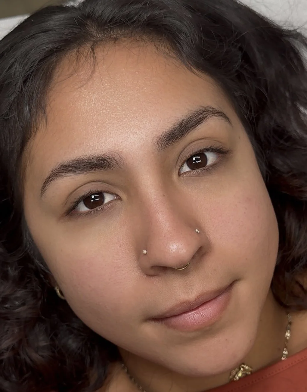 Close-up of a woman with wavy dark hair, brown eyes, piercings on her nose, and a gentle smile.