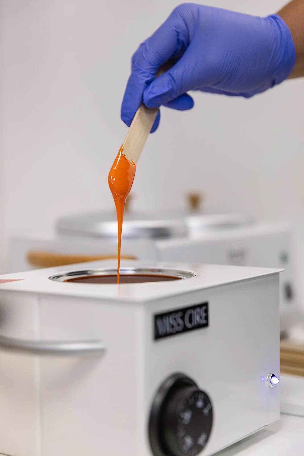 A gloved hand holding a wooden stick with orange wax dripping into a white melting pot labeled 'MISS CIRE'.