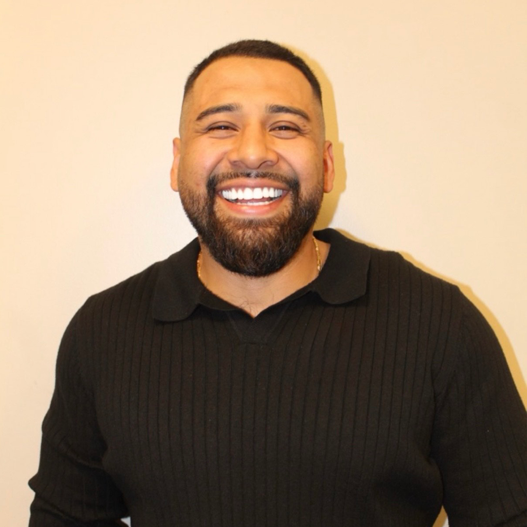 A smiling man with a beard and short hair, wearing a black ribbed polo shirt, stands against a plain light-colored wall.