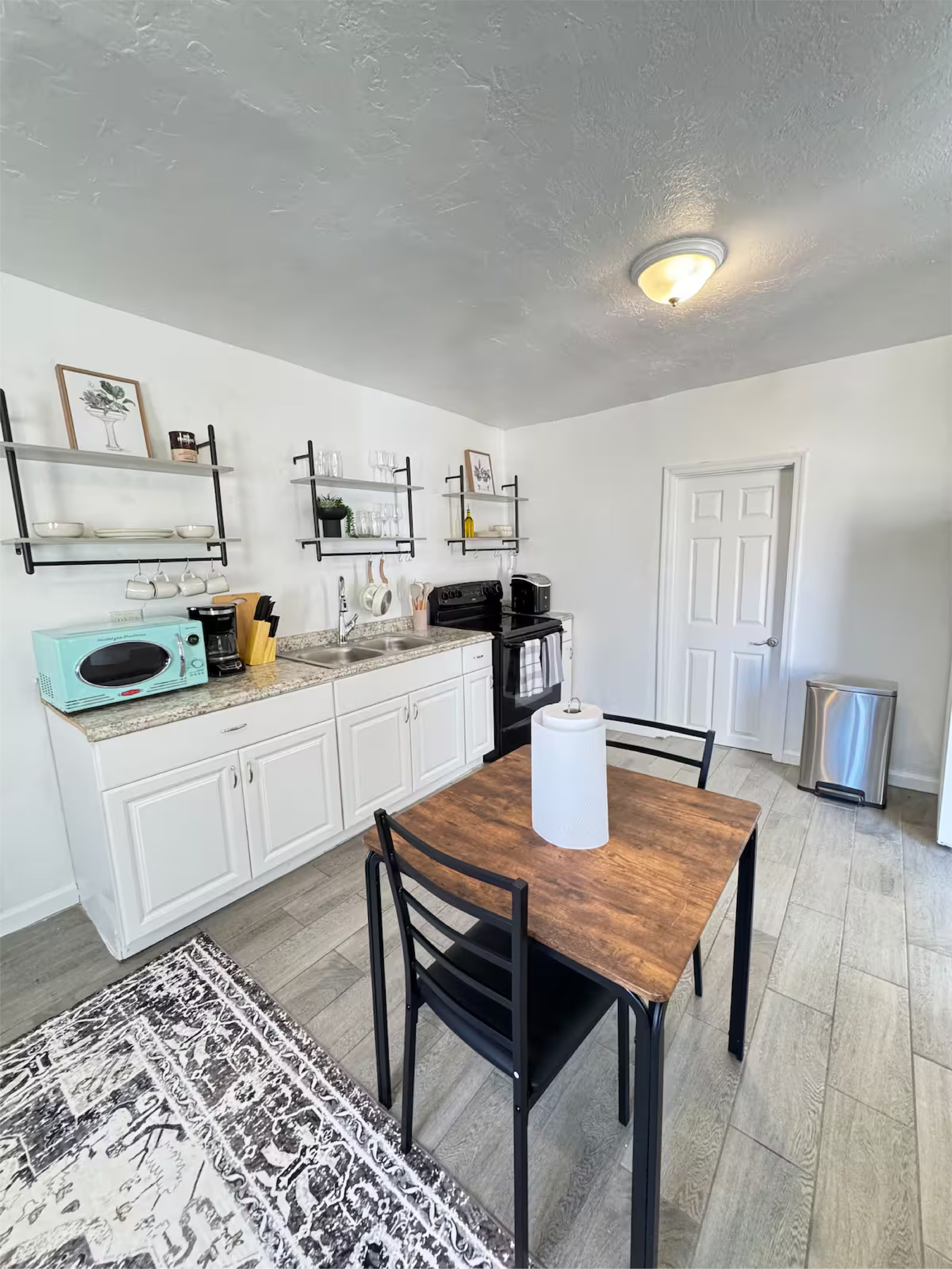 A kitchen with white cabinets, open shelves with glasses, bowls, framed art, and plants. A wooden dining table with black chairs, a roll of paper towels, a microwave, toaster, and coffee maker. A door, a trash bin, and a ceiling light fixture.