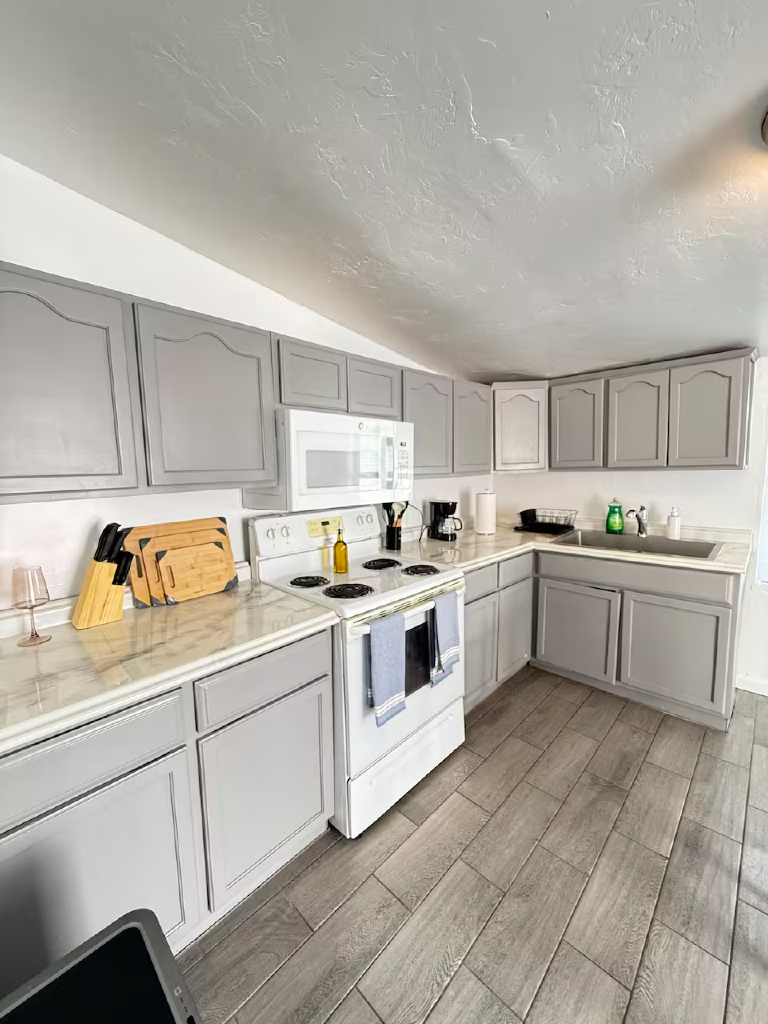 A kitchen with light gray cabinets, white countertops, and a white stove with a microwave above. There are knives and cutting boards on the counter, along with a glass of wine, oil bottle, coffee maker, paper towel, dish soap, and sink with a faucet. The floor has large, tiled, wood-like planks. The ceiling is textured, and a ceiling light is visible.