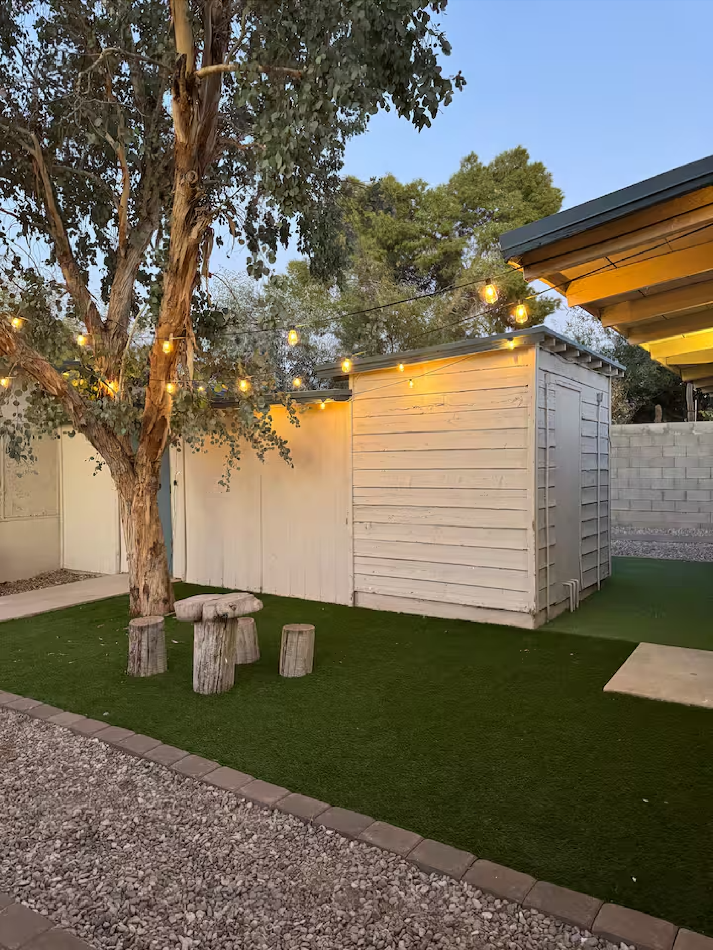 A backyard scene with a large tree, string lights hanging above, and a small white shed. There are four wooden stools arranged around the tree on artificial grass, and a gravel pathway border in the foreground.