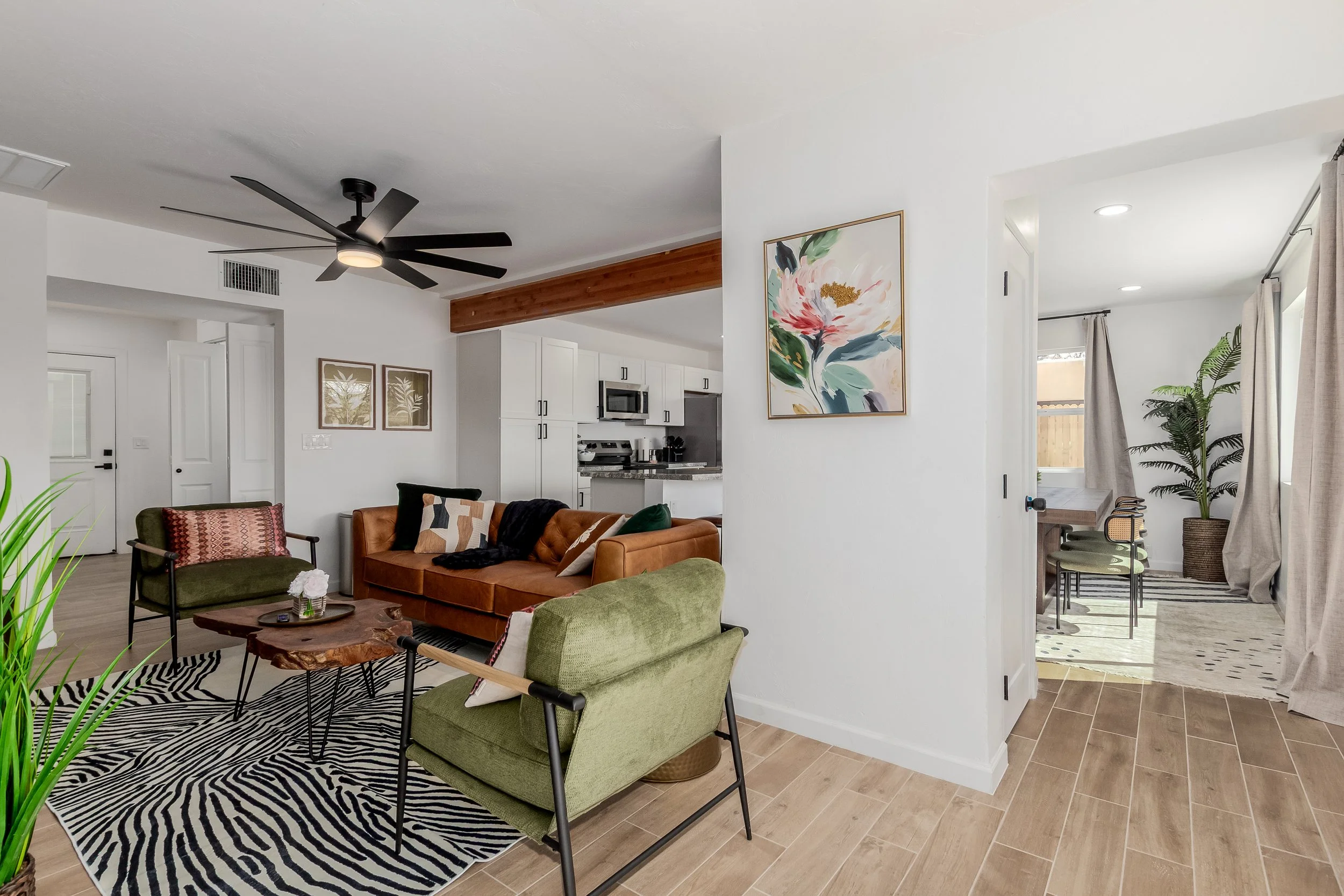 Living room with green and brown chairs, a brown sofa, a wooden coffee table, black and white zebra print rug, white walls with artwork, ceiling fan, open kitchen with white cabinets, and adjacent dining area with large windows and curtains.