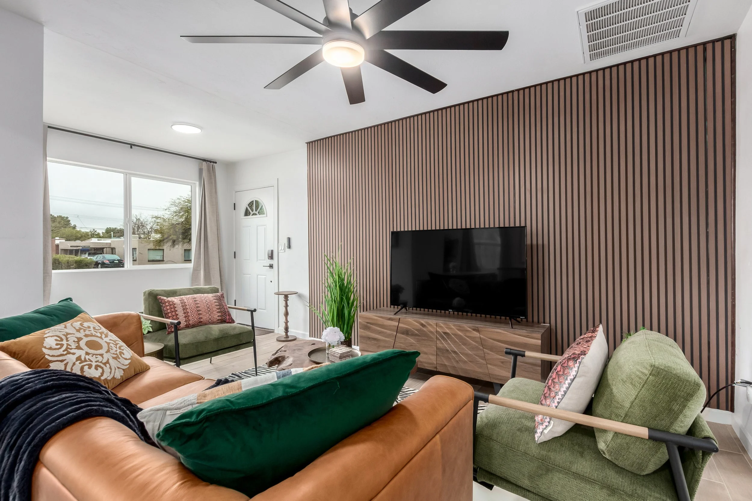 Living room with a ceiling fan, a flat-screen TV on a wooden stand, a leather sofa with cushions, multiple armchairs, a side table, a large window with curtains, and a decorative plant.