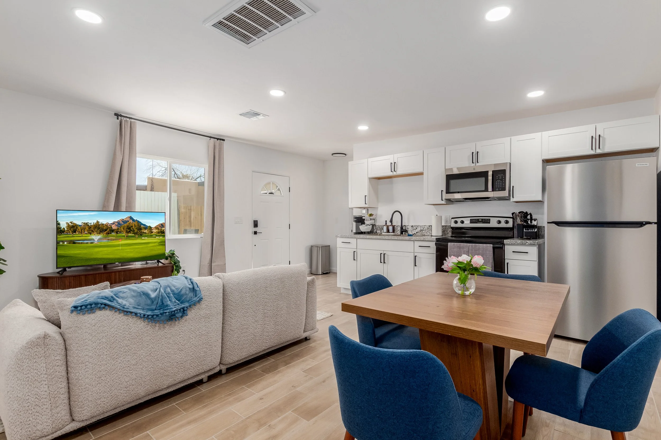 Open-concept living room and kitchen with white walls, beige sectional sofa with a blue blanket, wooden dining table with blue chairs, kitchen with white cabinets, stainless steel appliances, and small window with curtains.