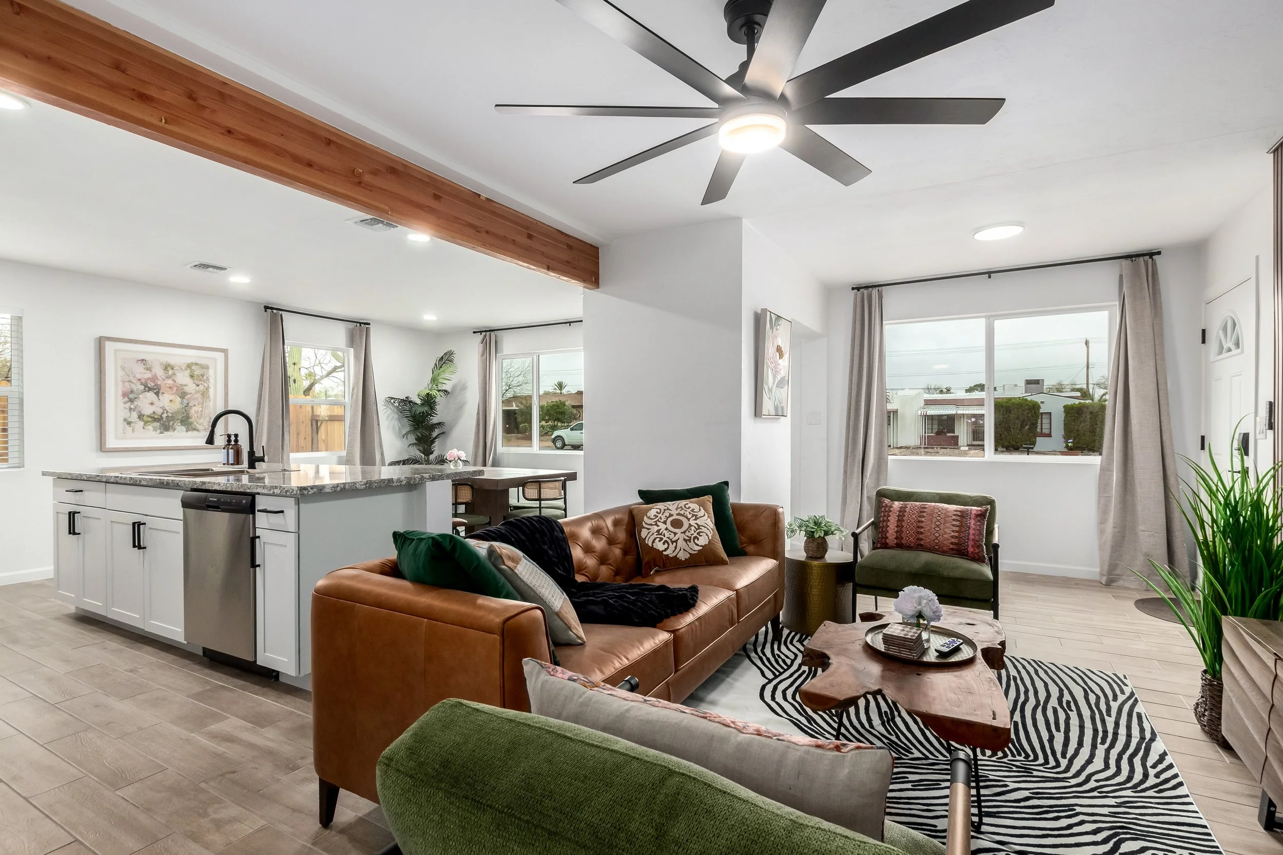 Modern living room with brown leather sofa, green and patterned armchairs, coffee table, area rug, large windows, ceiling fan, open kitchen with white cabinets and granite countertops, dining area, houseplants, and artwork on the walls.