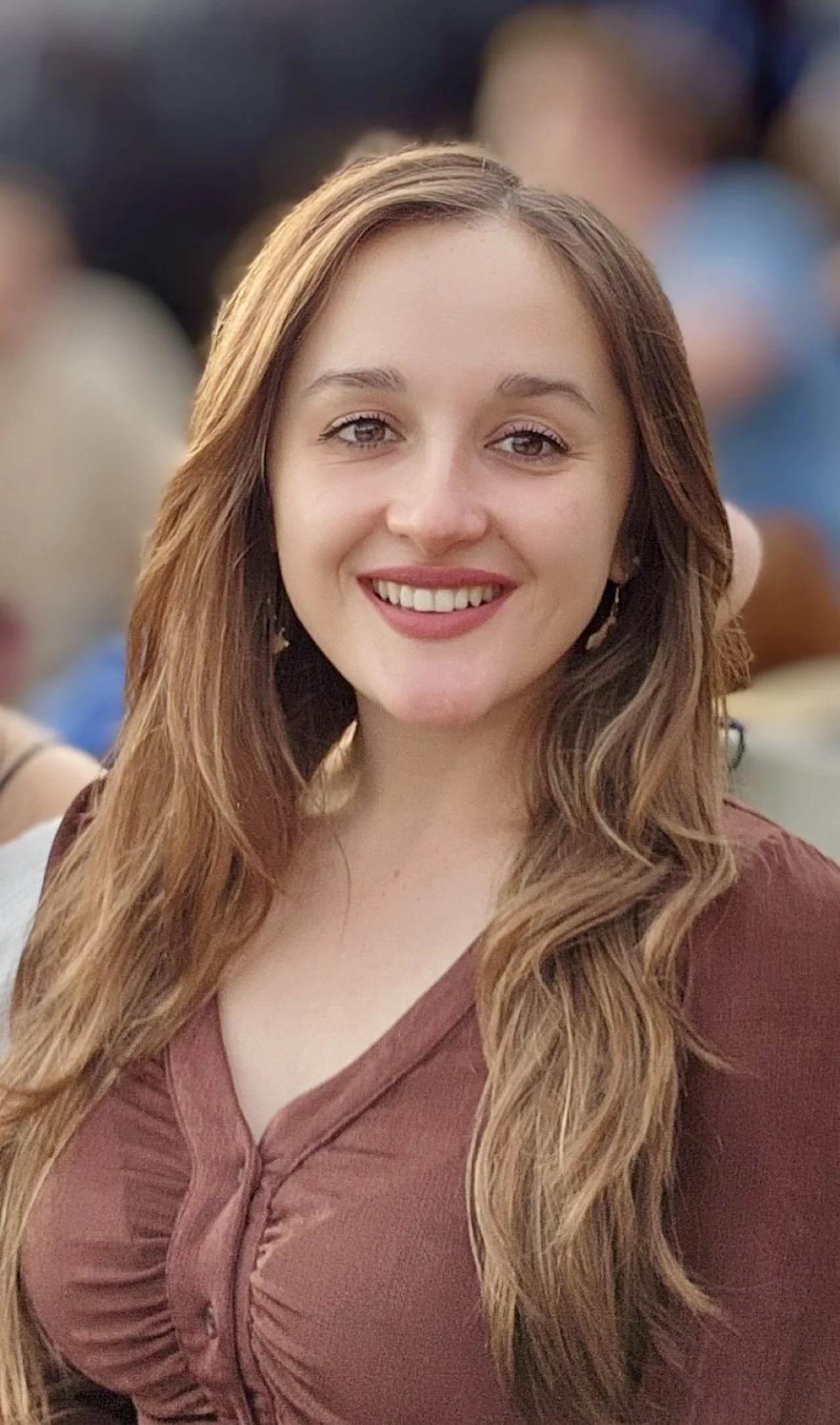 Close-up of a smiling young woman with long, wavy, light brown hair, wearing a rust-colored blouse.