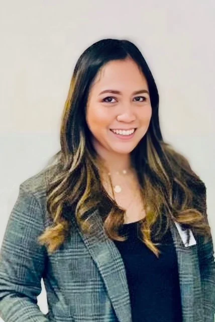A woman with long wavy hair, wearing a gray plaid blazer over a black top, smiling against a plain light background.