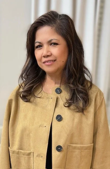 A woman with shoulder-length dark hair, wearing a tan blazer with large black buttons and a black top underneath, standing in front of a neutral background.