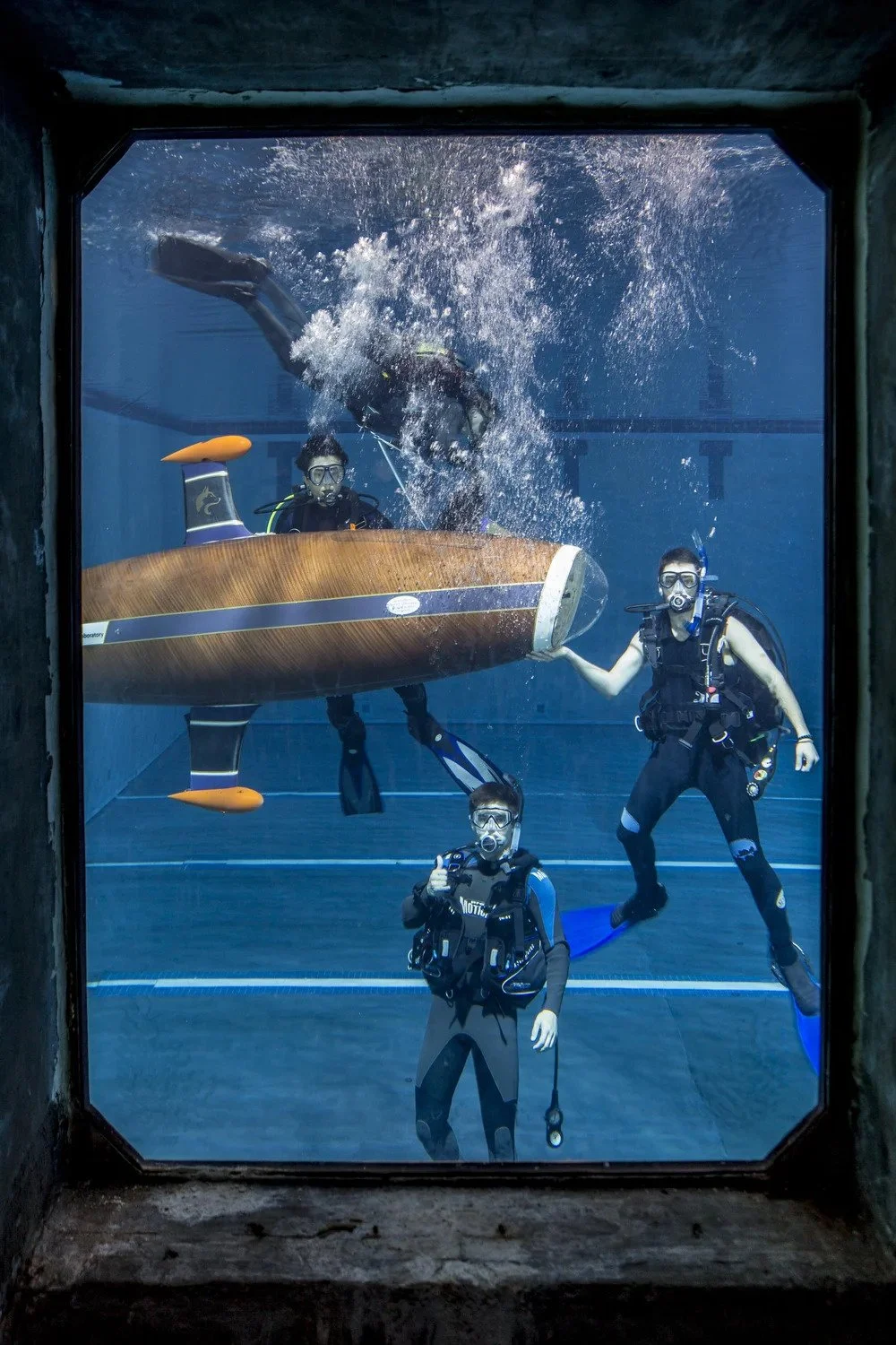 Three scuba divers underwater submerged with a wooden submarine, one in the background swimming above, the others near it, one holding the boat model, all wearing masks and gear.