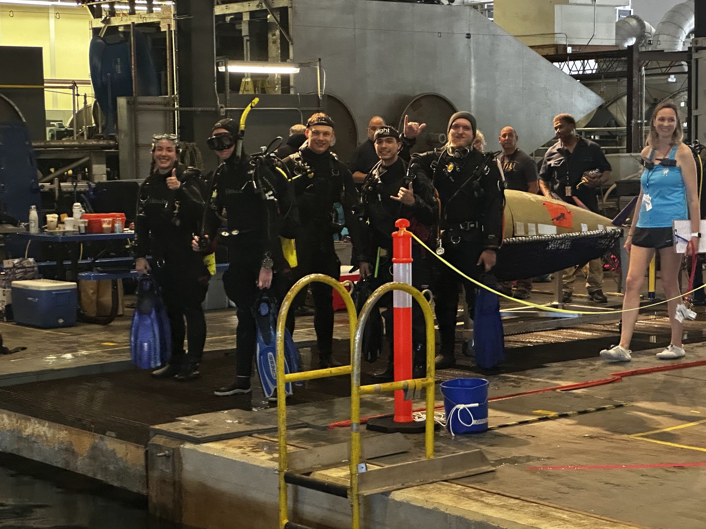 A group of six people in scuba diving gear standing on a dock, smiling, with a submarine in the background. They are holding dive equipment and appear to be preparing for a dive.
