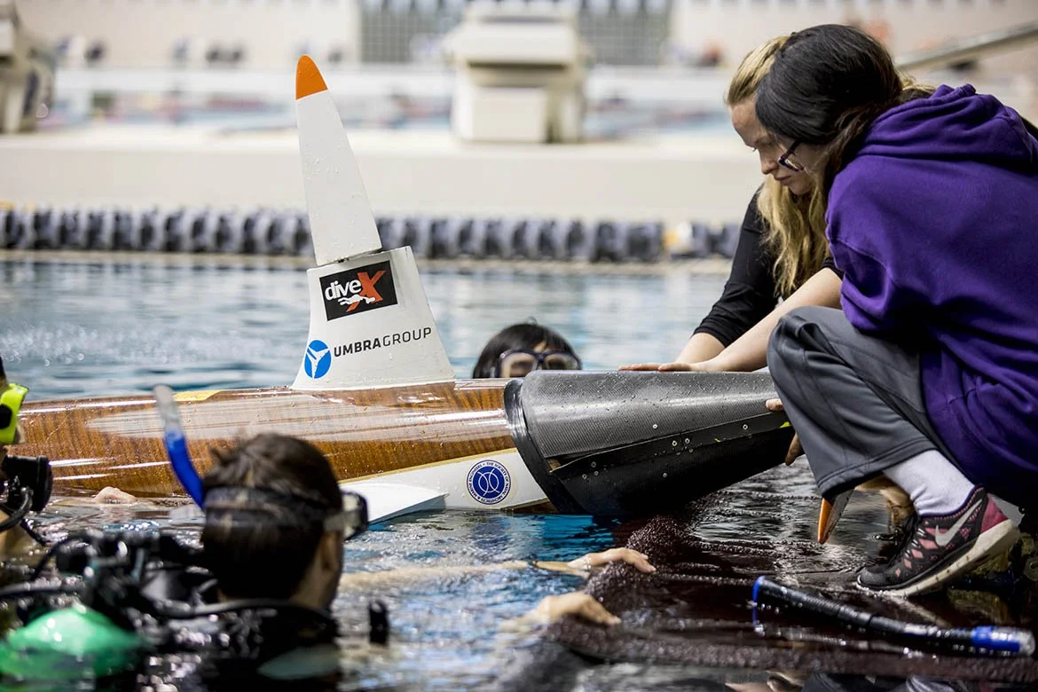 A group of people working on a submerged human-powered submarine in a swimming pool, with one person helping another at the front of the vessel, and a person in diving gear near the side.