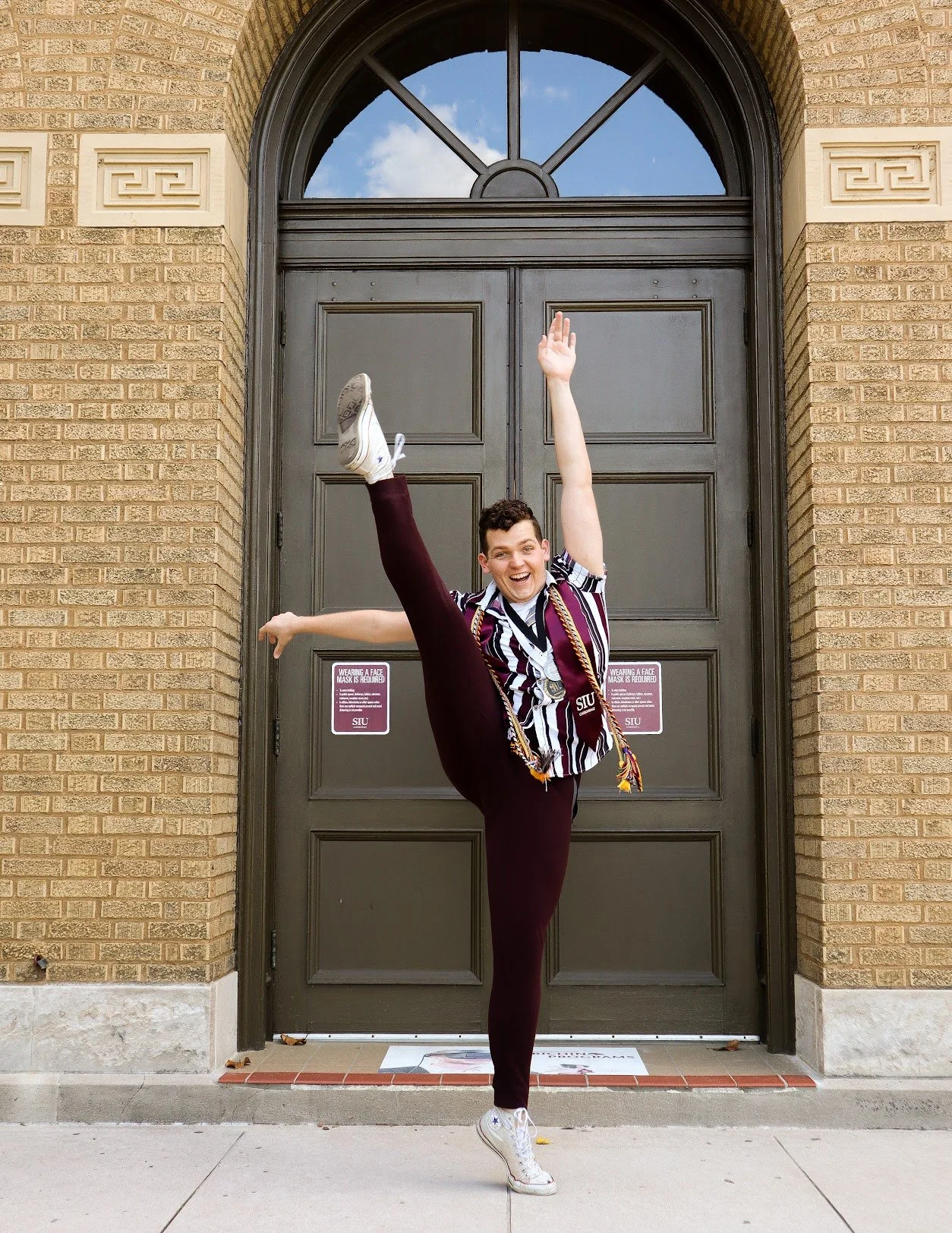 A young person performing a high kick in front of a large, dark-colored door with a brick building facade, wearing a striped shirt, maroon pants, and white sneakers, smiling excitedly.