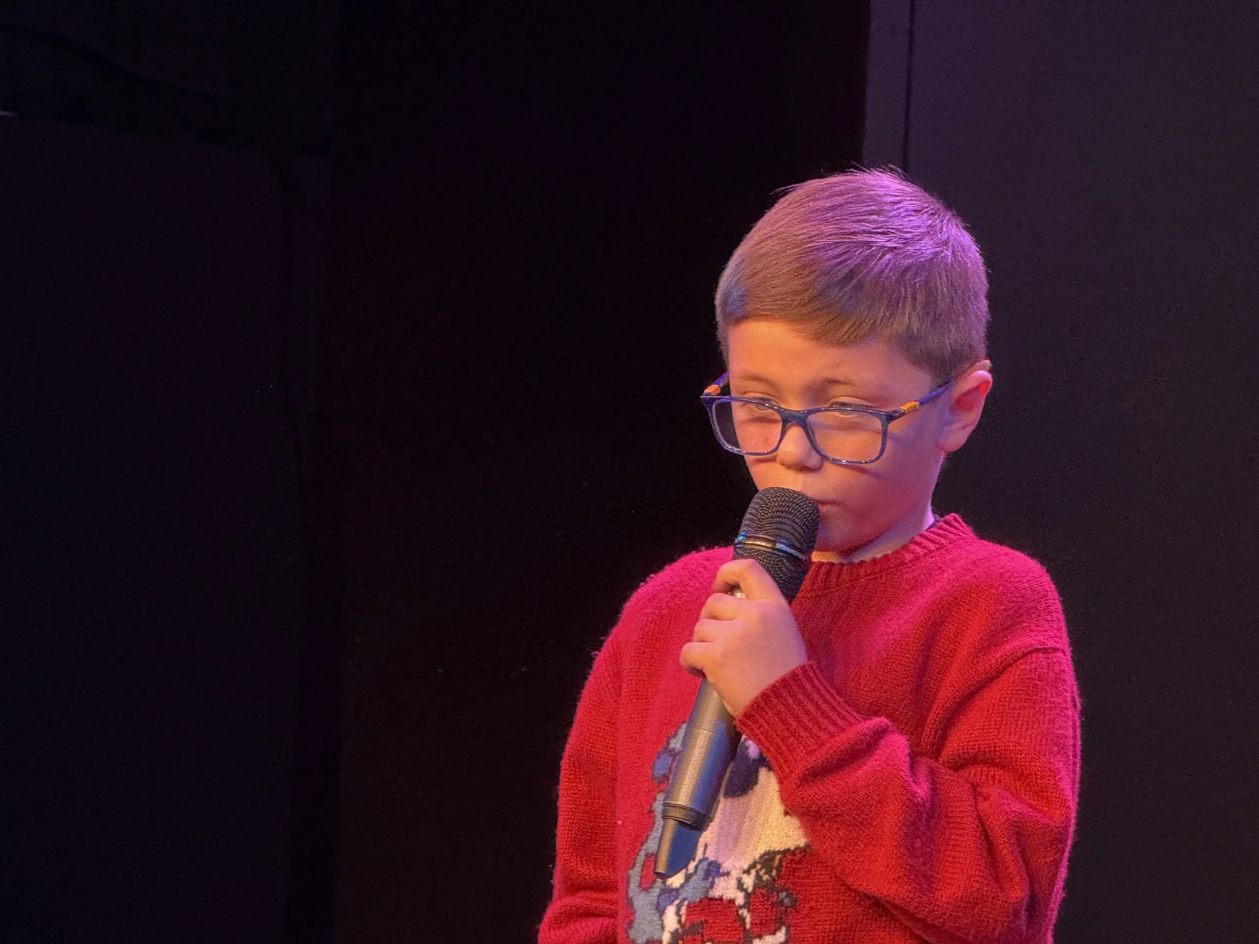 A young boy with glasses and light brown hair, wearing a red sweater with a reindeer design, holding a microphone and speaking or singing into it against a dark background.