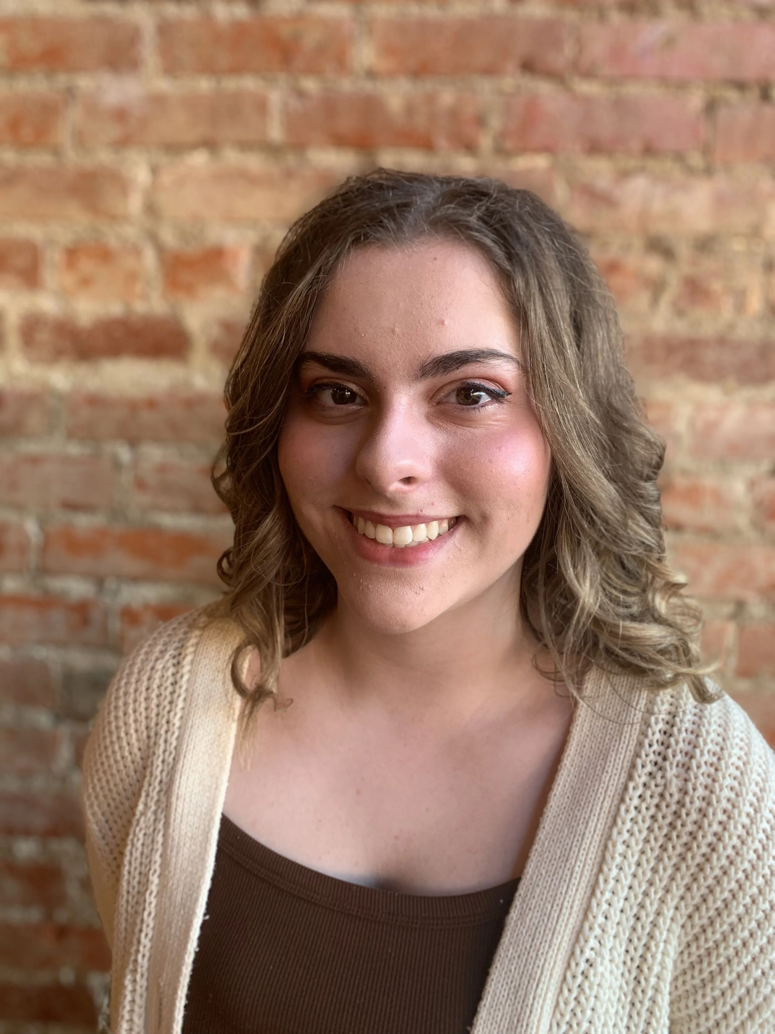 A young woman with curly brown hair and a light complexion, smiling, wearing a beige knit cardigan over a brown top, against a brick wall background.
