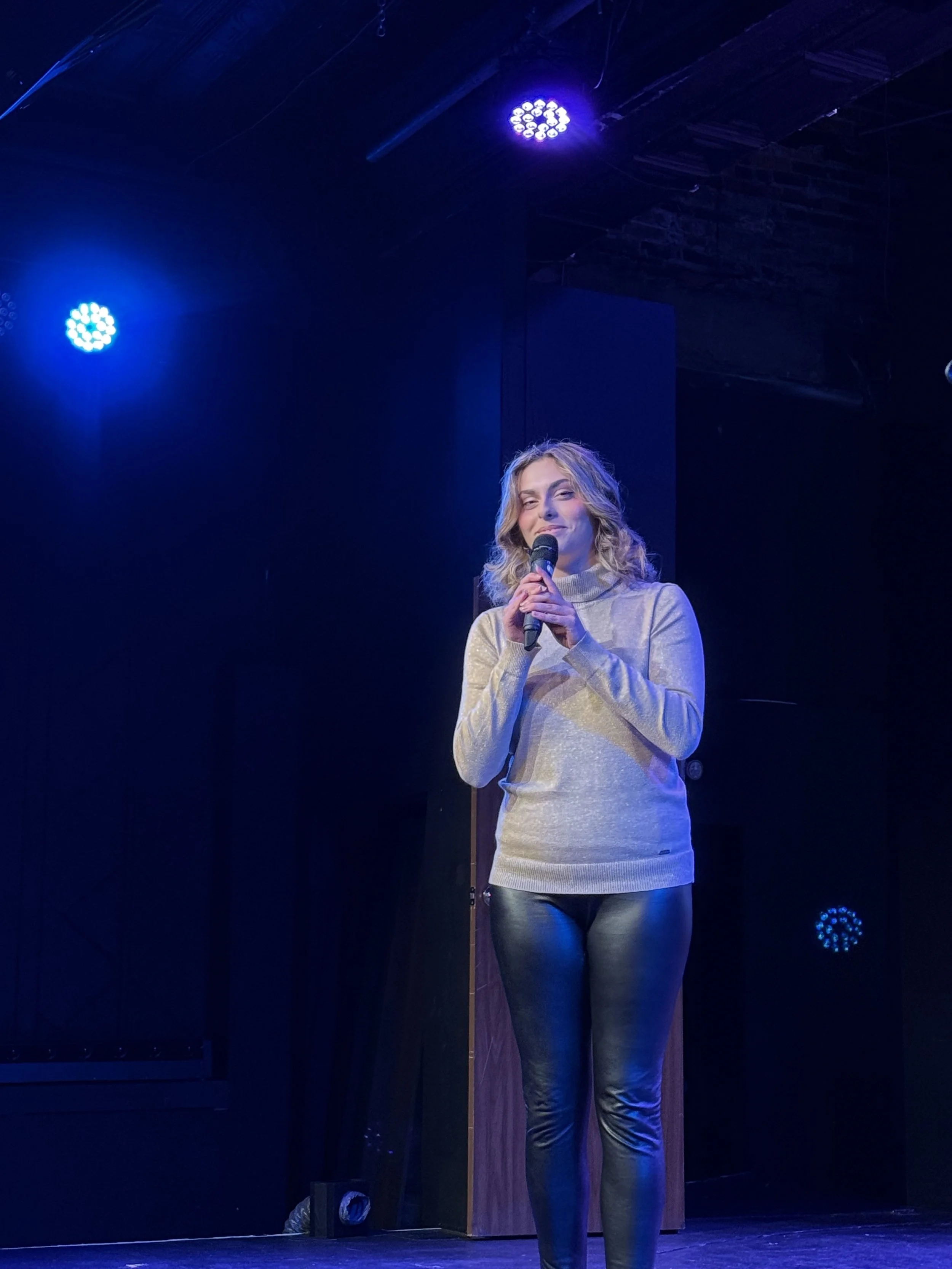 A woman stands on a stage holding a microphone, illuminated by bluish stage lighting, wearing a gray sweater and black leather pants, with a dark background.