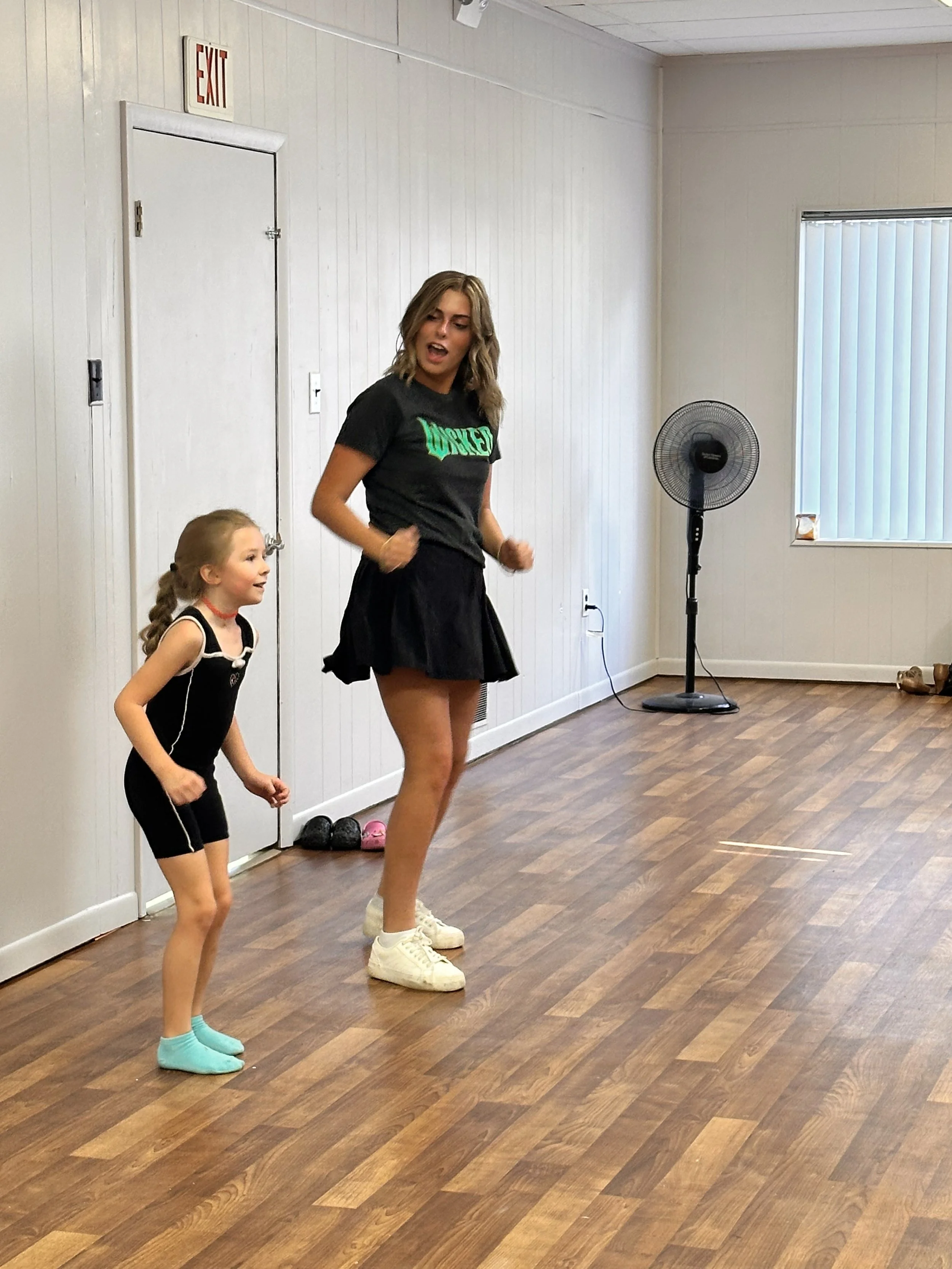 A woman and a young girl dancing in a room with wooden floors, a fan, and a window with vertical blinds.