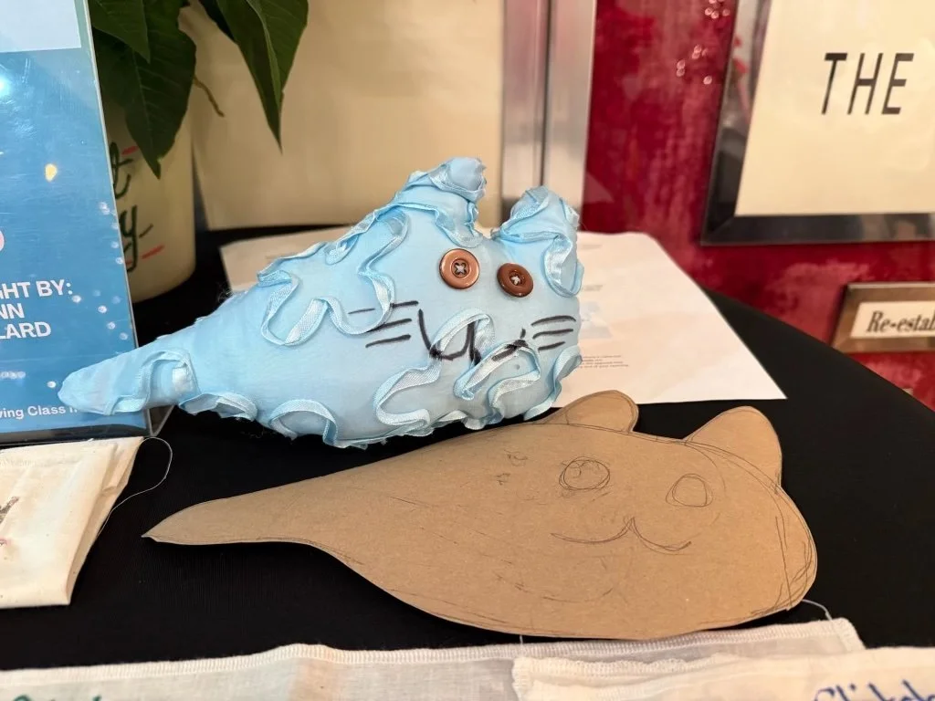 A homemade craft of a cat made from blue fabric with ruffles, button eyes, and drawn facial features, placed next to a paper sketch of a similar cat design on a black surface.