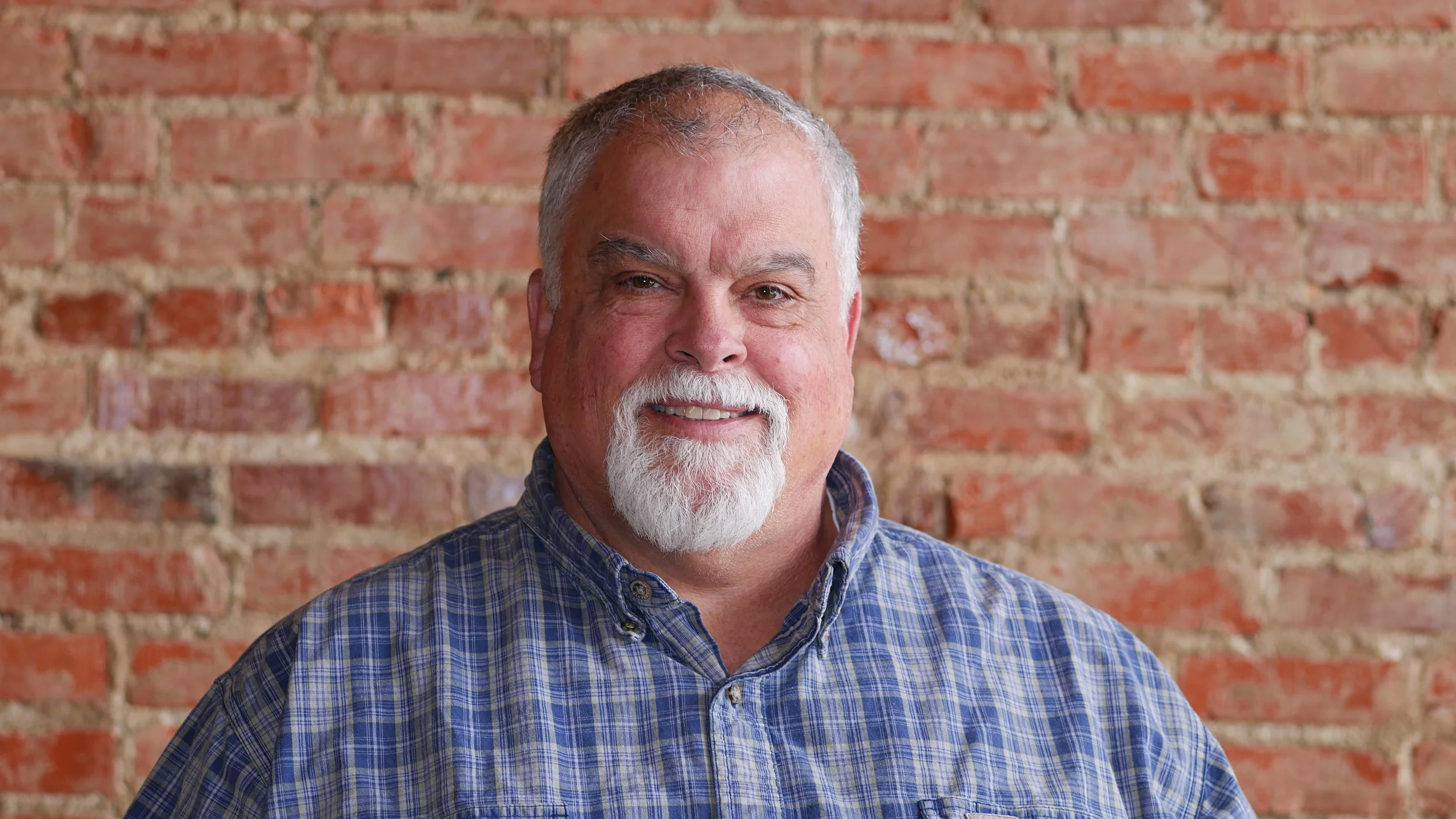 A smiling middle-aged man with a white beard and gray hair, wearing a blue checkered shirt, standing in front of a red brick wall.
