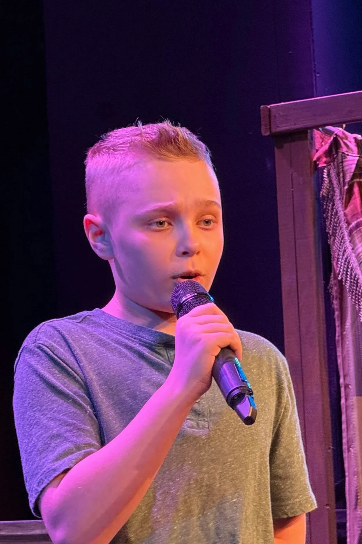 A young boy holding a microphone, standing on a stage with purple lighting, wearing a gray t-shirt, with a serious or focused expression.