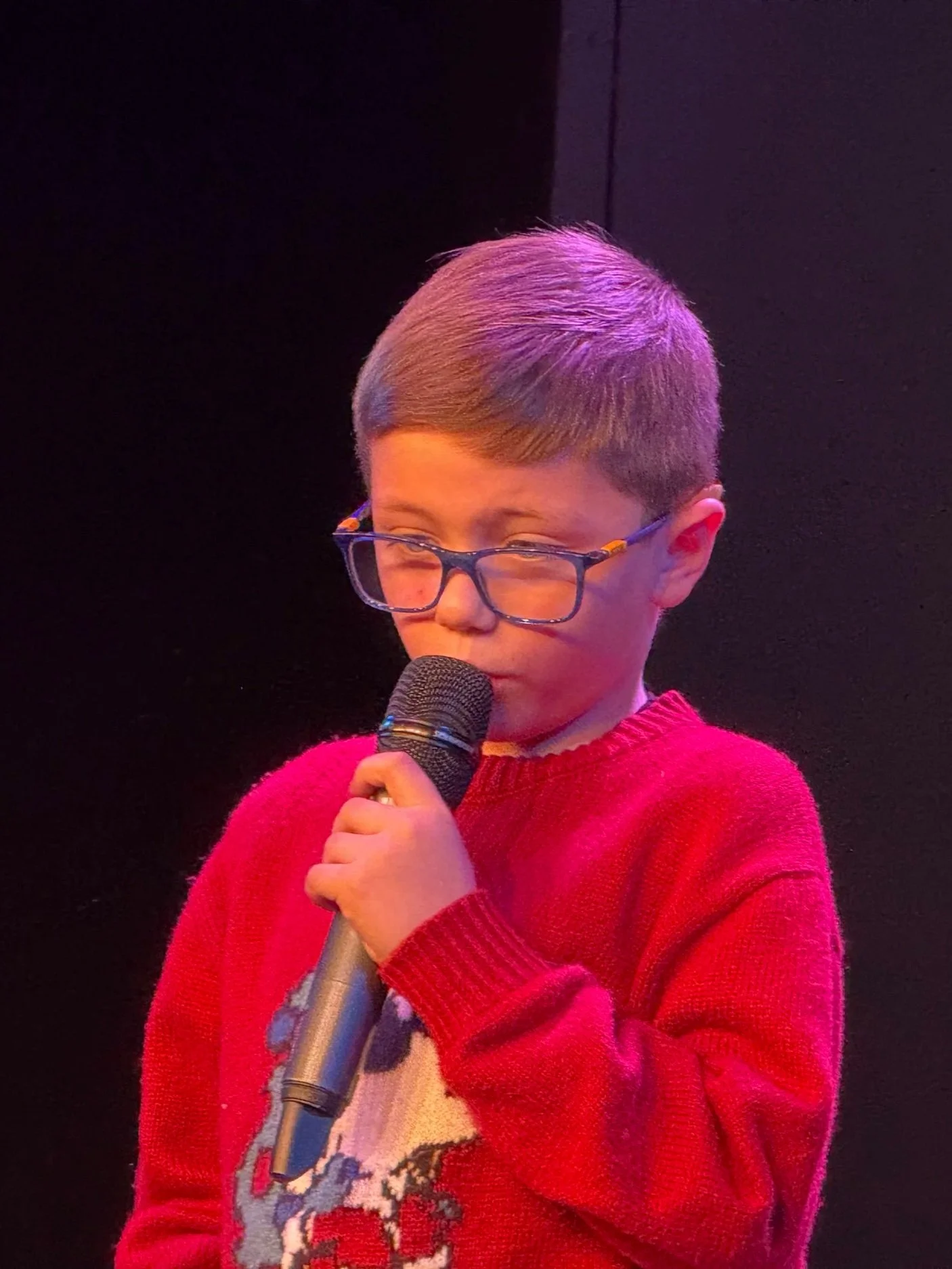 A young boy with glasses and short hair, wearing a red sweater, is holding a microphone and appears to be speaking or singing on stage.
