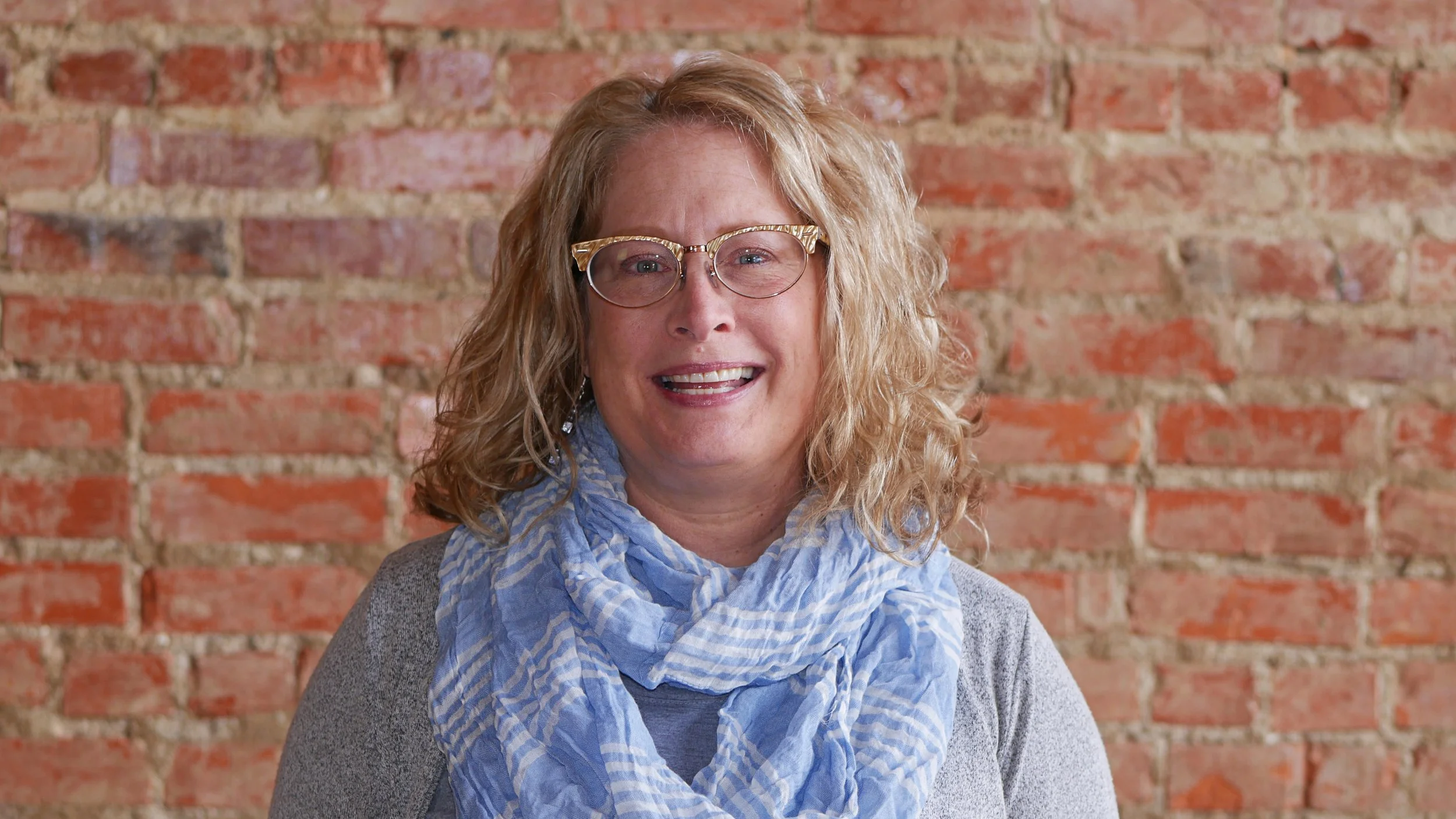 A woman with curly blonde hair, glasses, and a blue scarf stands in front of a brick wall.