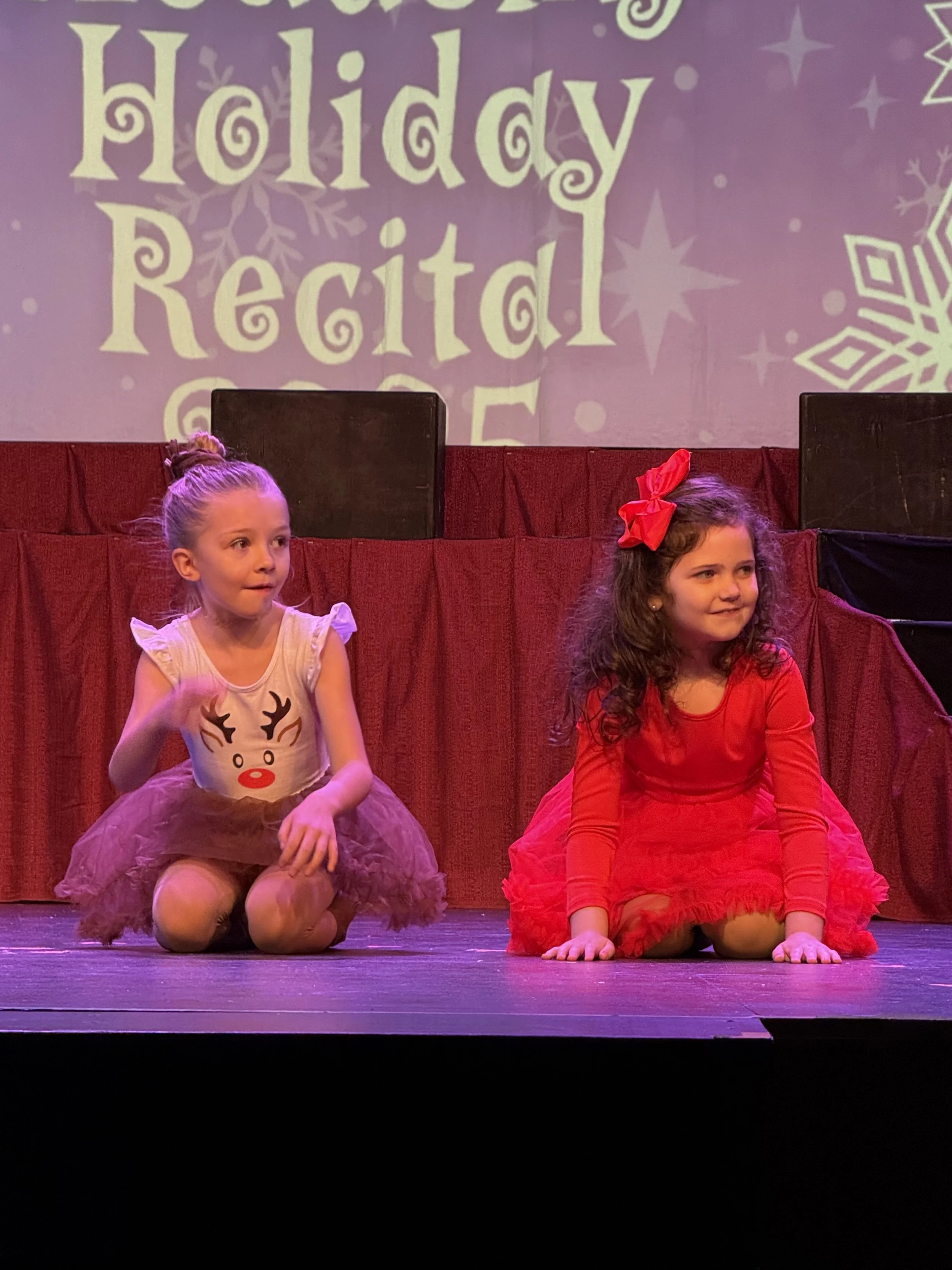 Two young girls sitting on the stage floor during a holiday Recital, with a holiday-themed backdrop and stage decorations.
