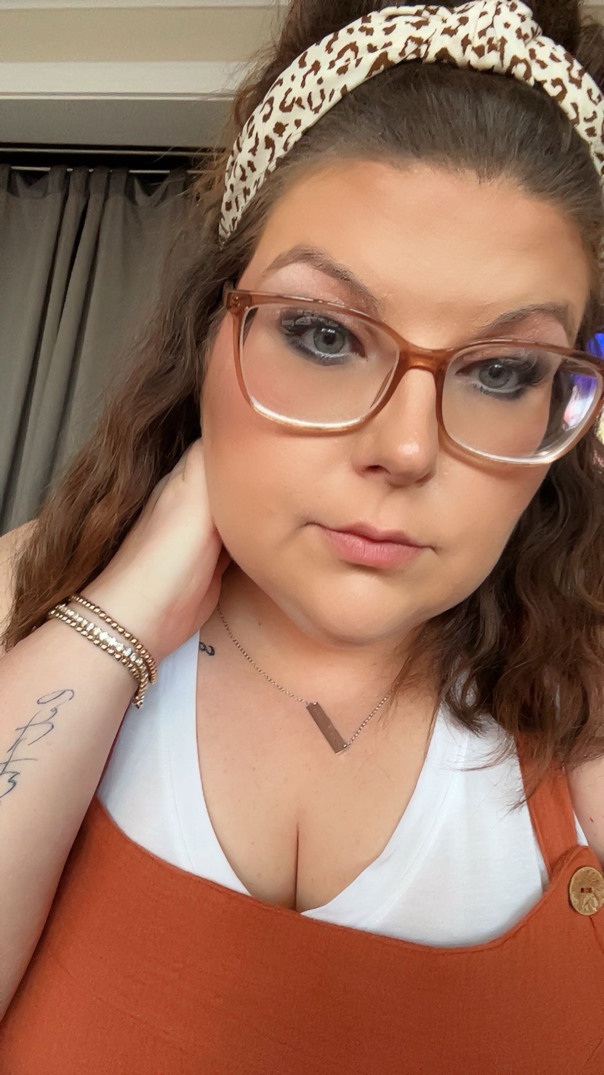 A young woman with curly brown hair wearing glasses, a white headband with animal print, a white shirt, and an orange sleeveless top. She has a tattoo on her left arm, bracelets on her left wrist, and a necklace with a brown, rectangular pendant. Her hand is resting on the back of her neck, and she appears to be indoors near a window with curtains.
