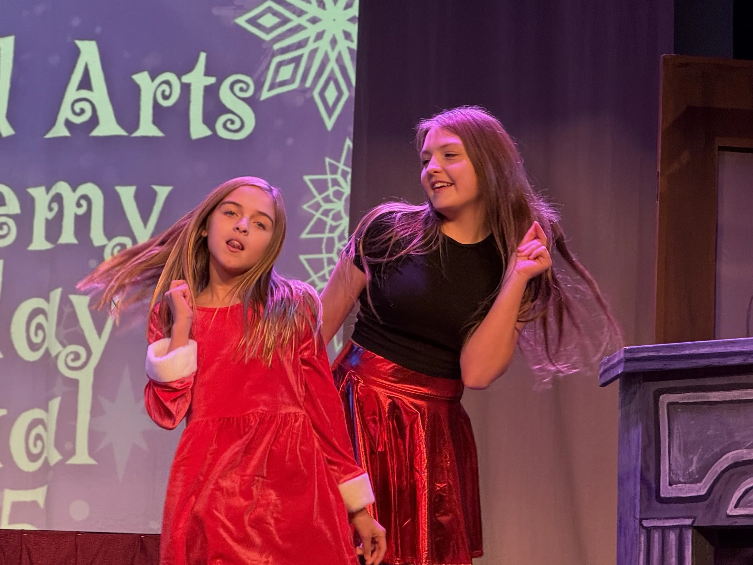 Two young girls performing on stage during a Christmas-themed event, with a festive backdrop reading 'Arts' and holiday designs.