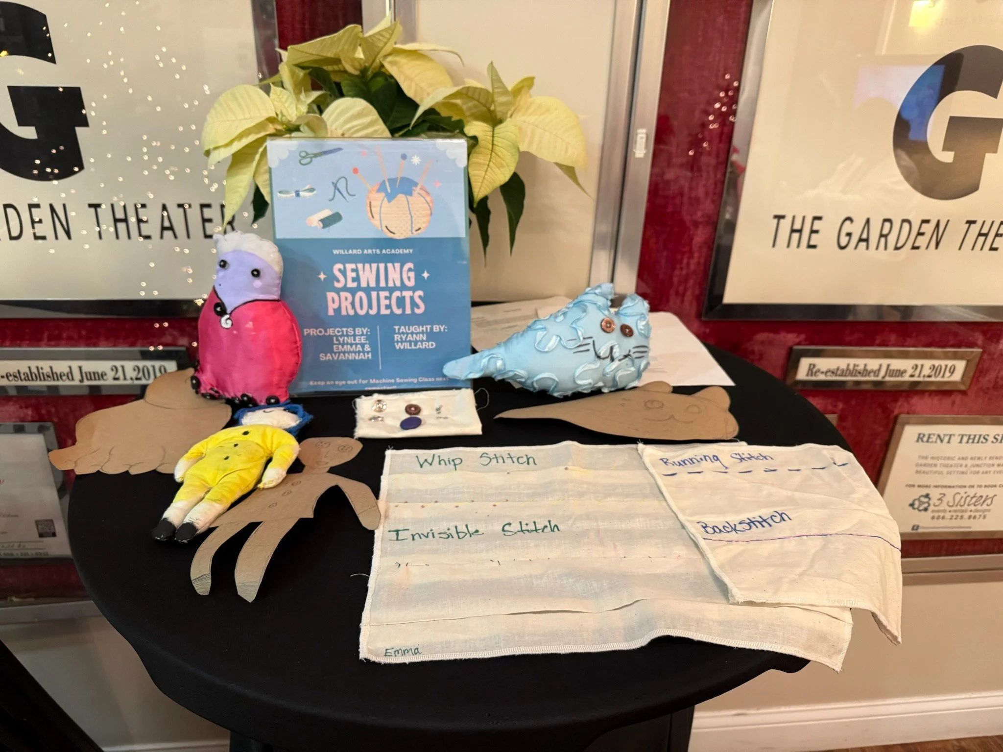 Display table with sewing projects, including felt animal finger puppets, fabric appliqué animals labeled with sewing techniques, and a sewing projects booklet, set in front of a window with signs about the Garden Theater.