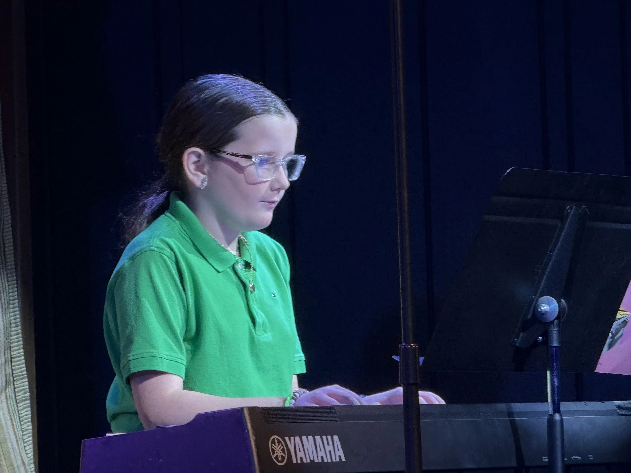 A young woman with glasses and earrings, wearing a green shirt, playing a Yamaha keyboard on stage with a dark background.