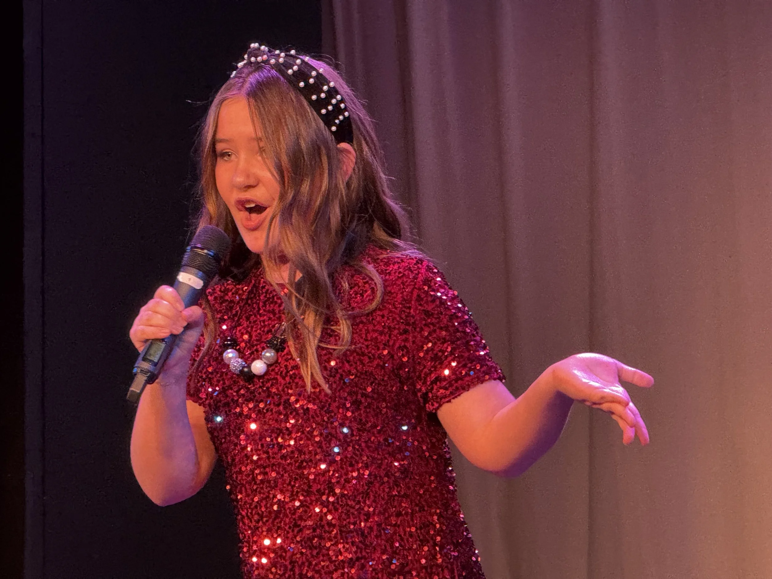 A woman singing into a microphone on stage, wearing a red, sequined dress and a pearl necklace, with a pearl headband in her hair.