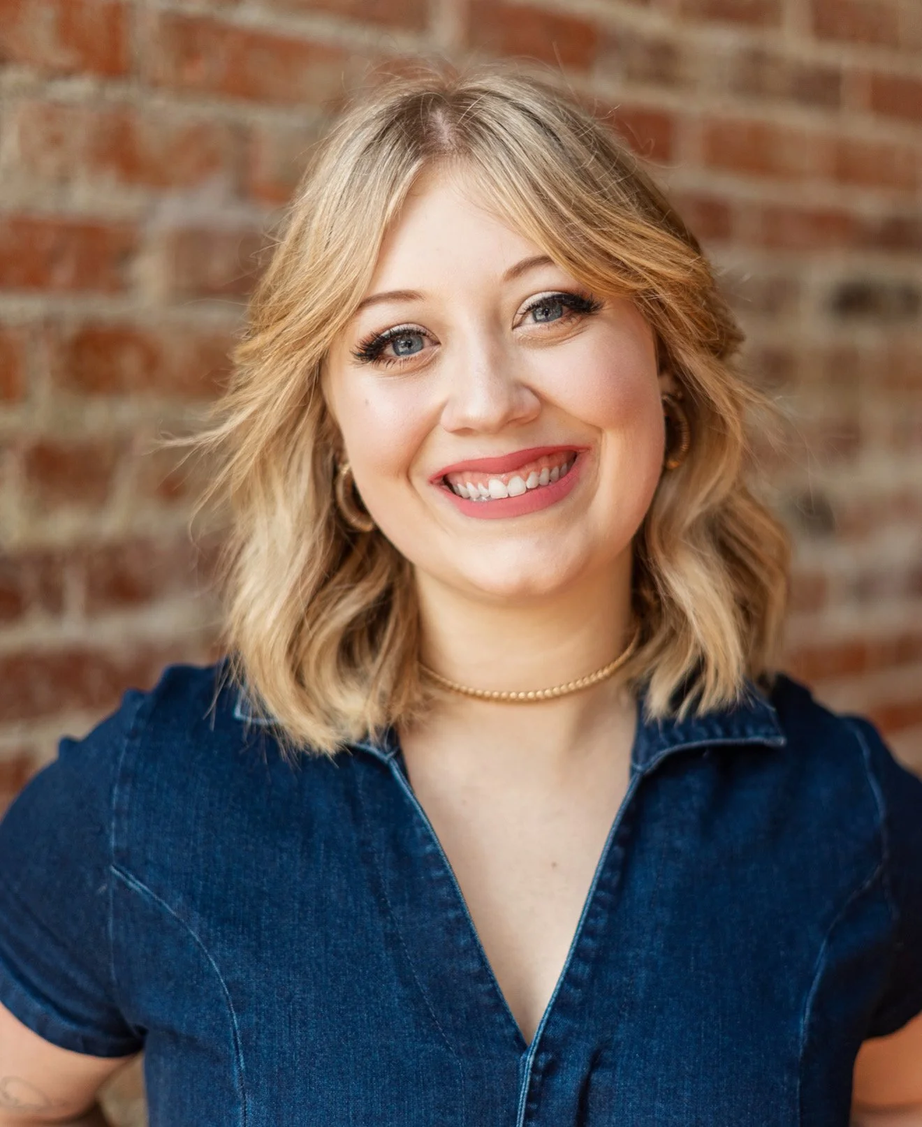 A smiling woman with blonde hair and light skin, wearing a dark denim shirt and a gold necklace, standing against a brick wall background.