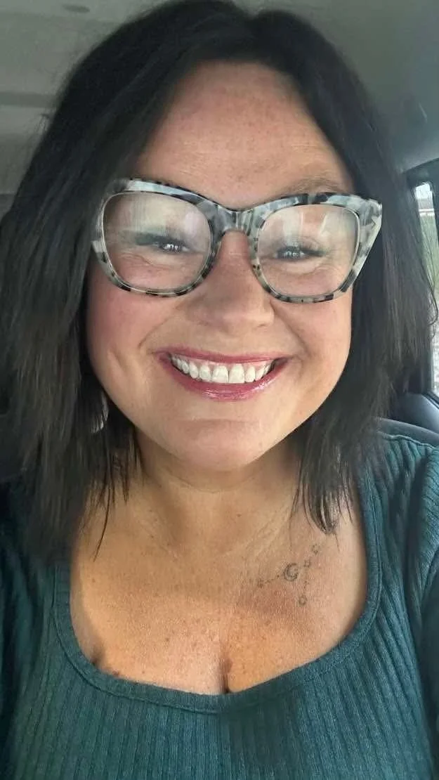 A smiling woman with shoulder-length dark hair, wearing large tortoiseshell glasses and a teal top, sitting inside a vehicle.