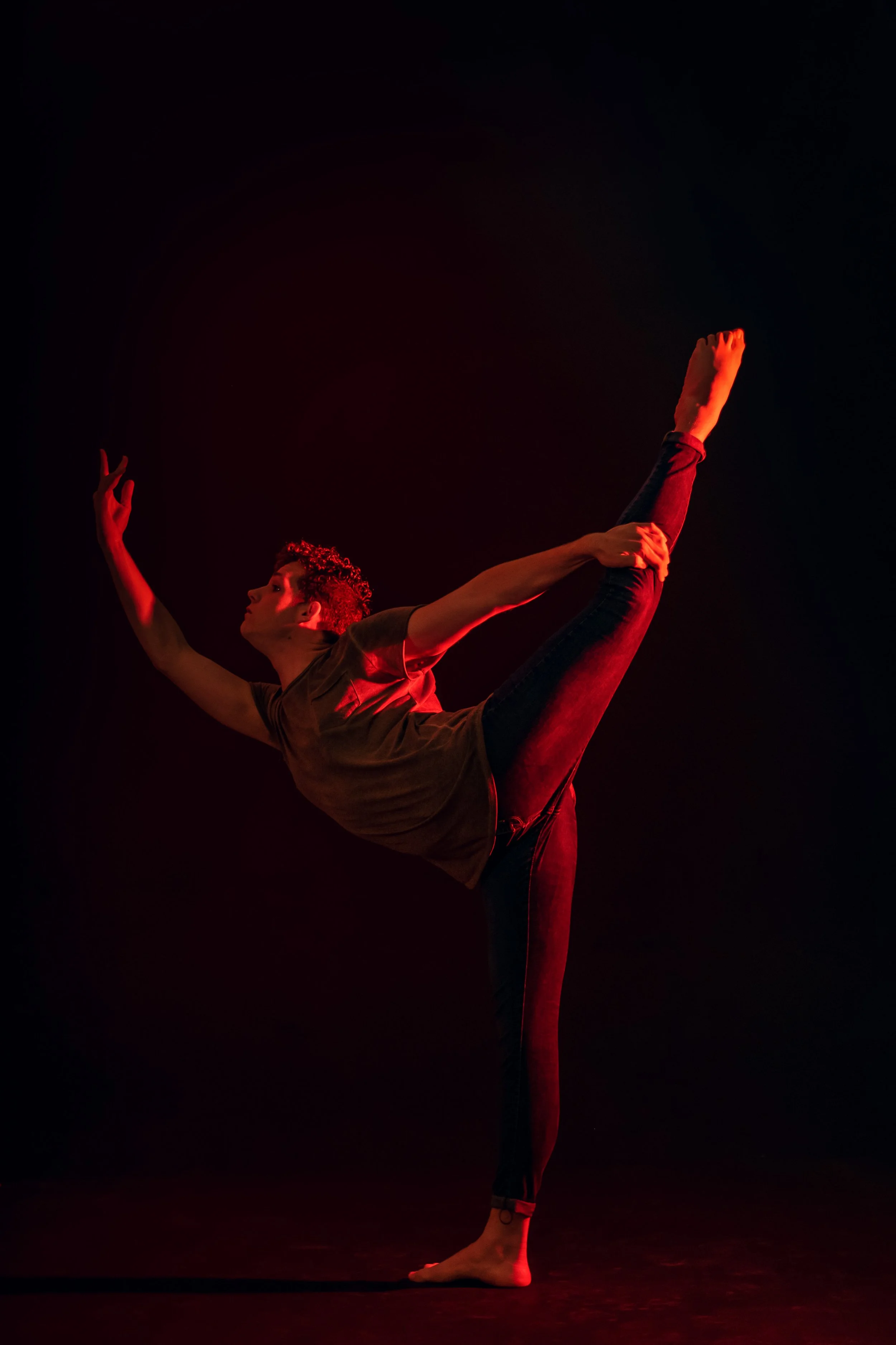 A dancer performing a standing split pose against a dark background, illuminated by red lighting.