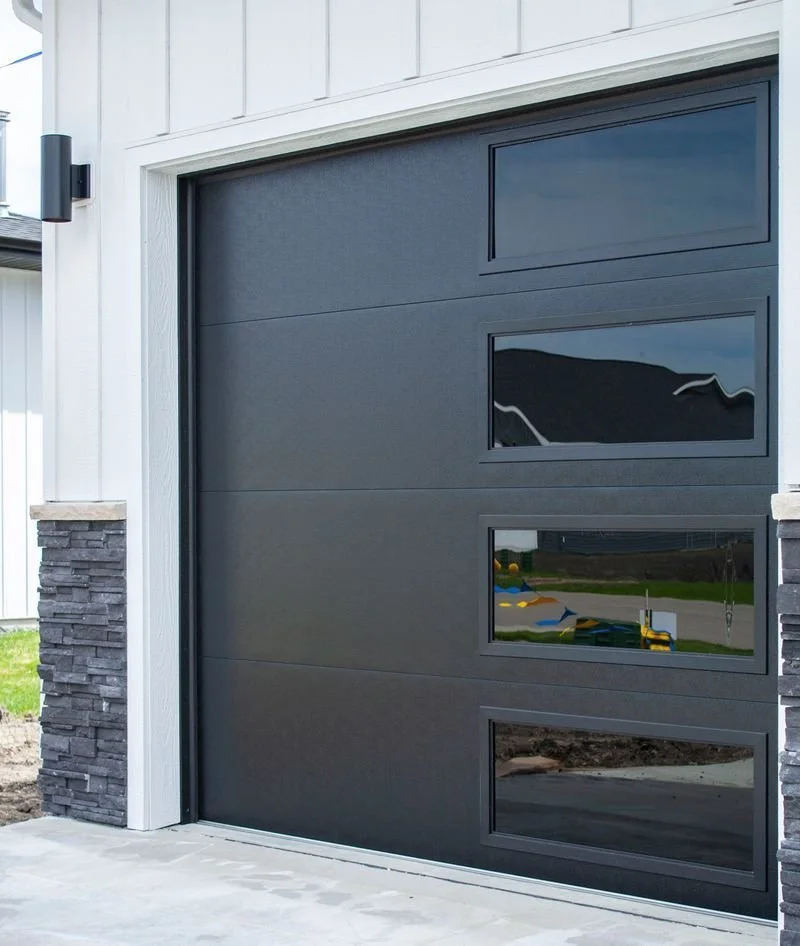 Modern black garage door with three rectangular windows, part of a white house with stone accents.