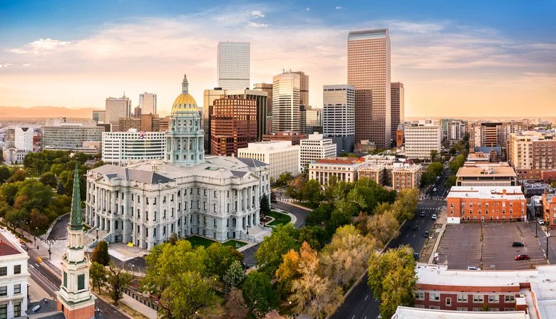 A city skyline featuring tall modern skyscrapers and a historic white government building with a gold dome, surrounded by trees and streets at sunset.
